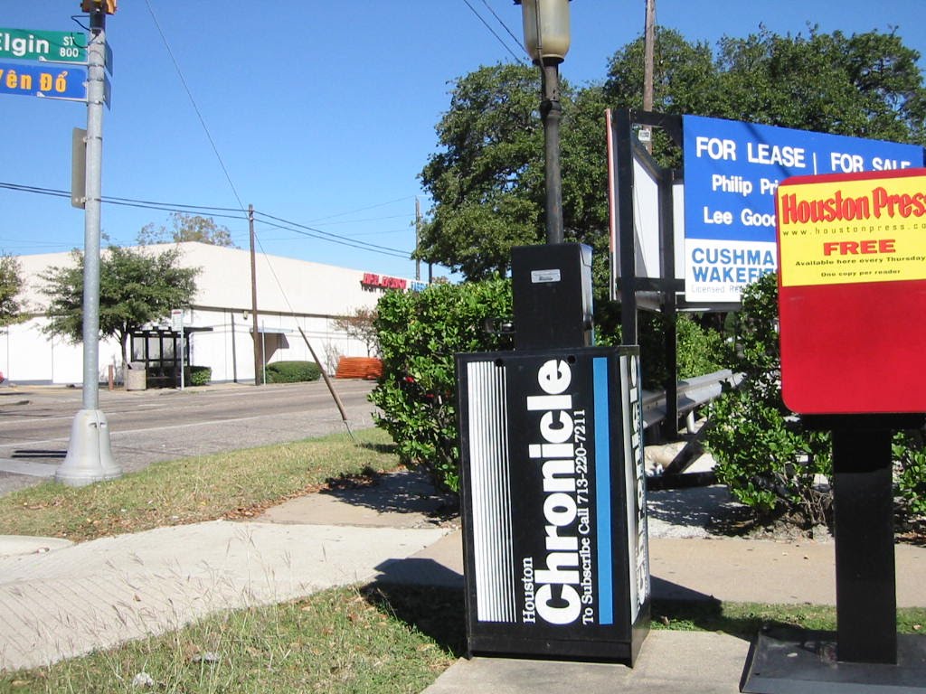 A newspaper stand displaying the Houston Chronicle on one side and a Houston Press box on the other, located at an intersection with street signs visible.