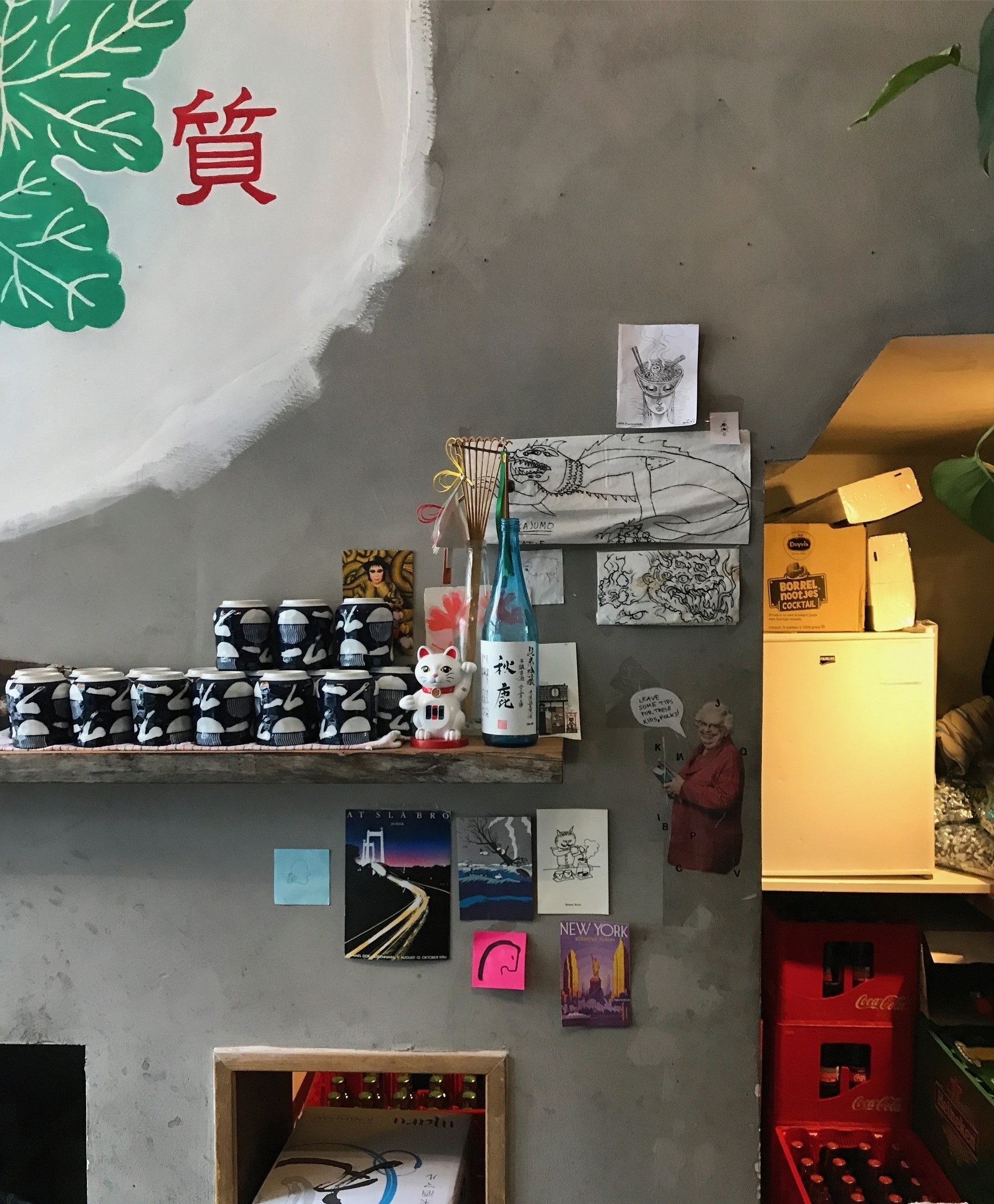 A gray wall displays stacked boxes, Japanese bottles, ceramic cups, and various posters with text.