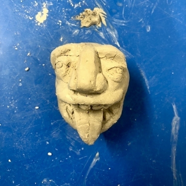 A small, gray clay sculpture of a face sits on blue background.