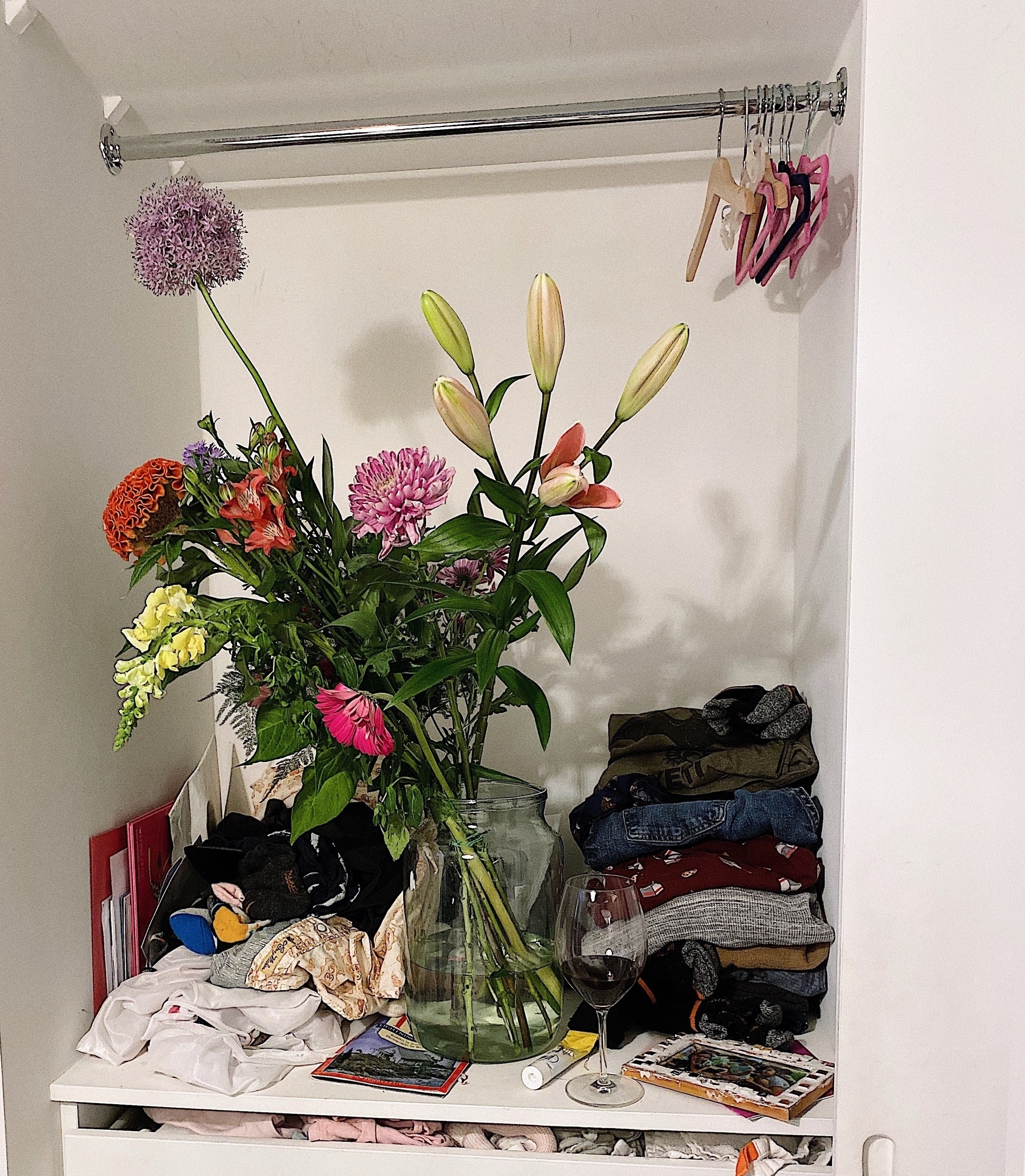 A white closet interior contains a vase of flowers, folded clothes, and a wine glass.