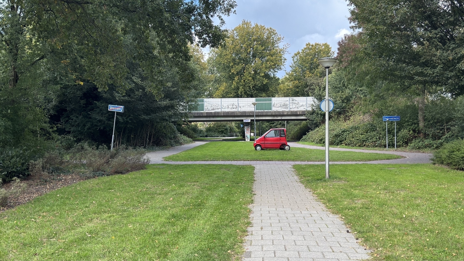 A red car is parked next to a paved path alongside a bridge with blue street signs.