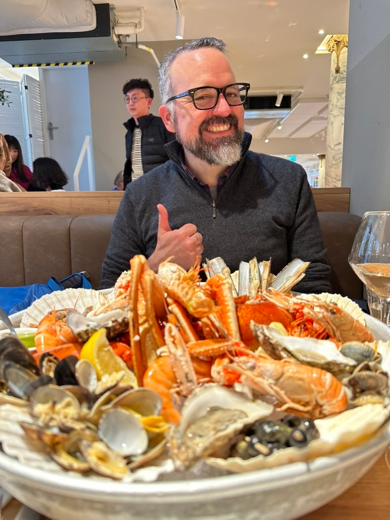 A man with a beard gestures towards a large seafood platter filled with crab, shellfish, and mussels.