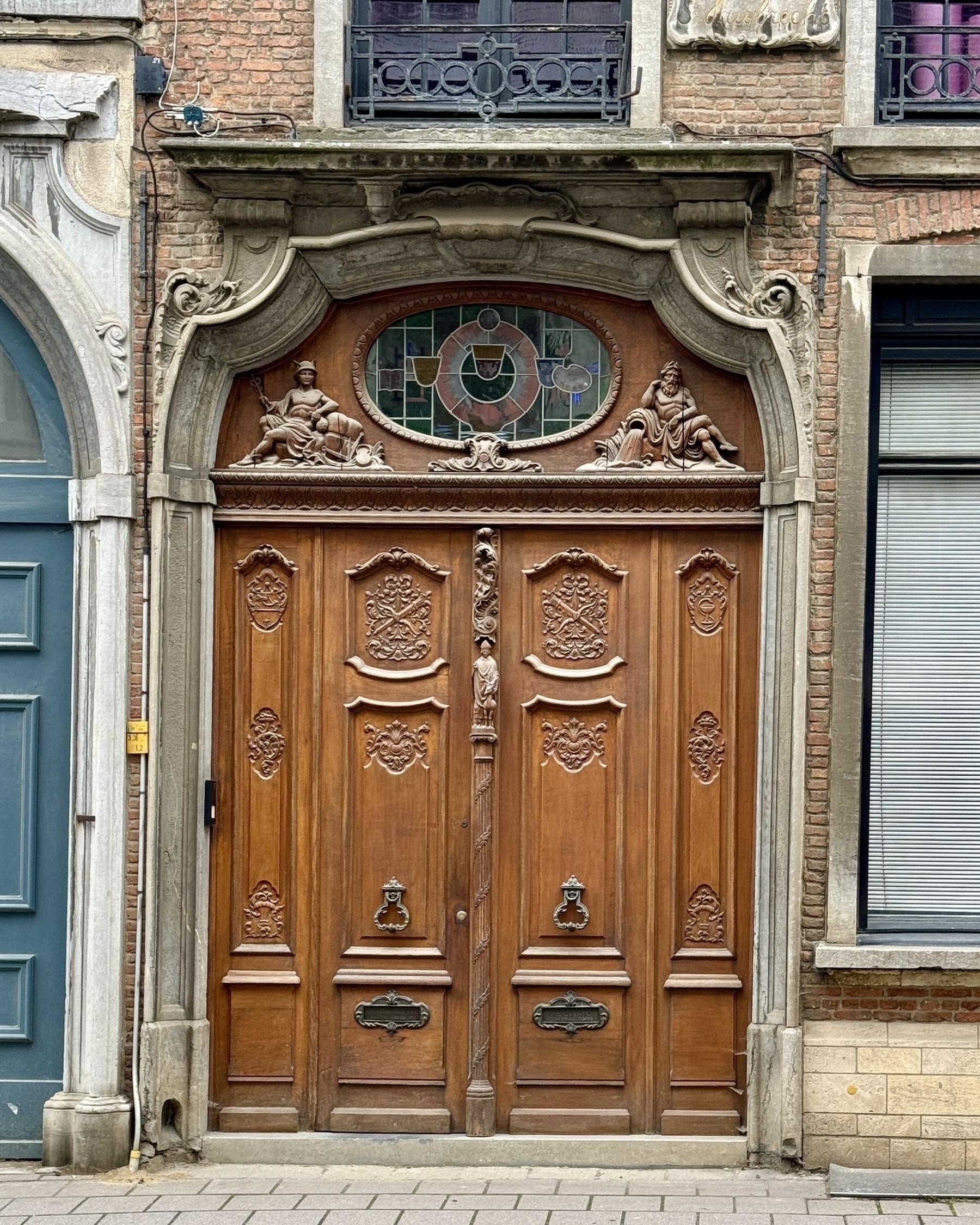 A detailed, ornate wooden door with sculpted figures and a stained-glass window is set into a brick facade.