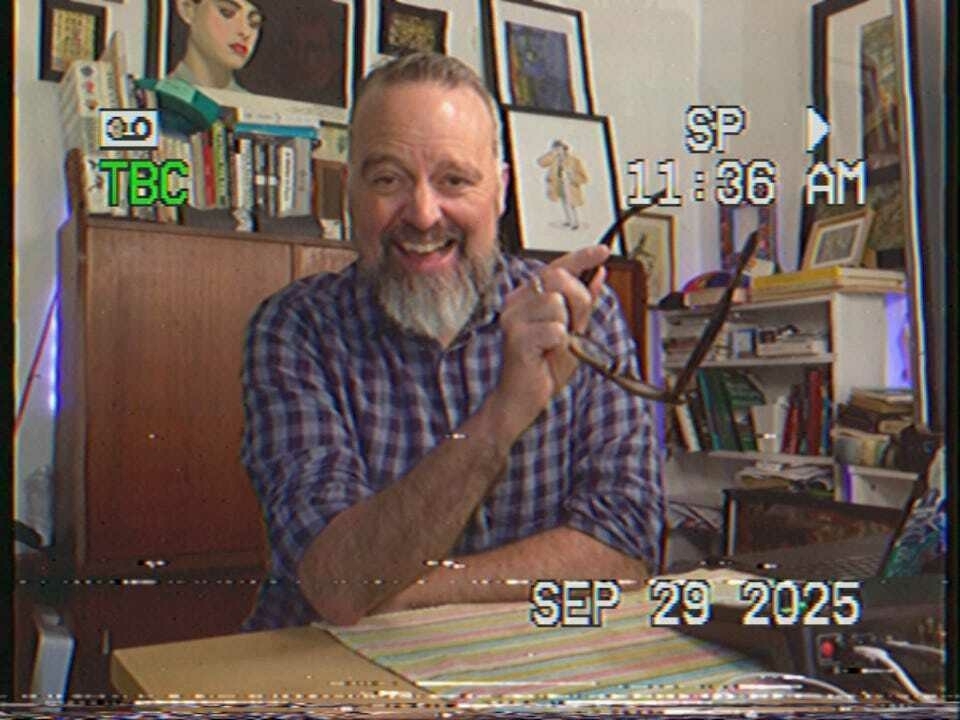 A man with gray hair is sitting at a table, sketching with a pencil, with text visible in the background.