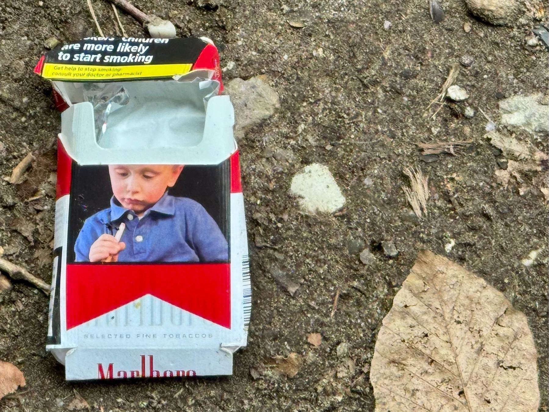 A discarded Marlboro cigarette pack with a child's image and "Selected Fine Tobacco" text is on the ground.