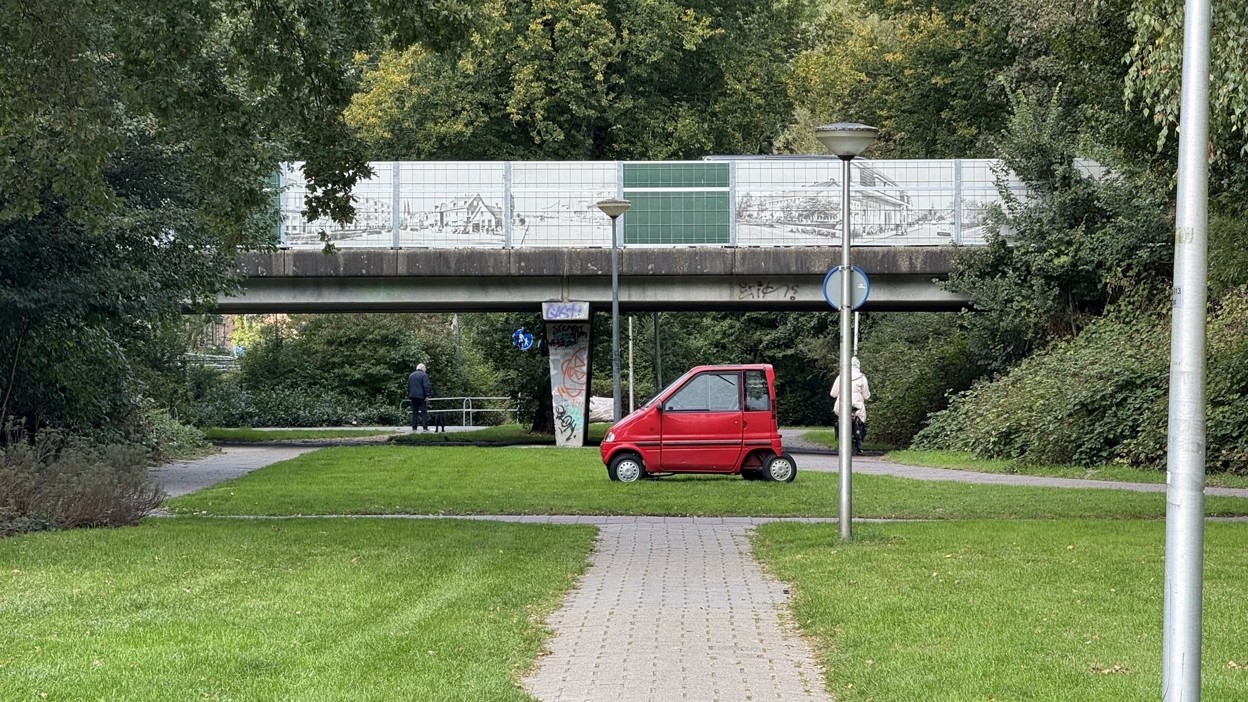A red mini-vehicle is parked on a paved path under a concrete bridge with graffiti.