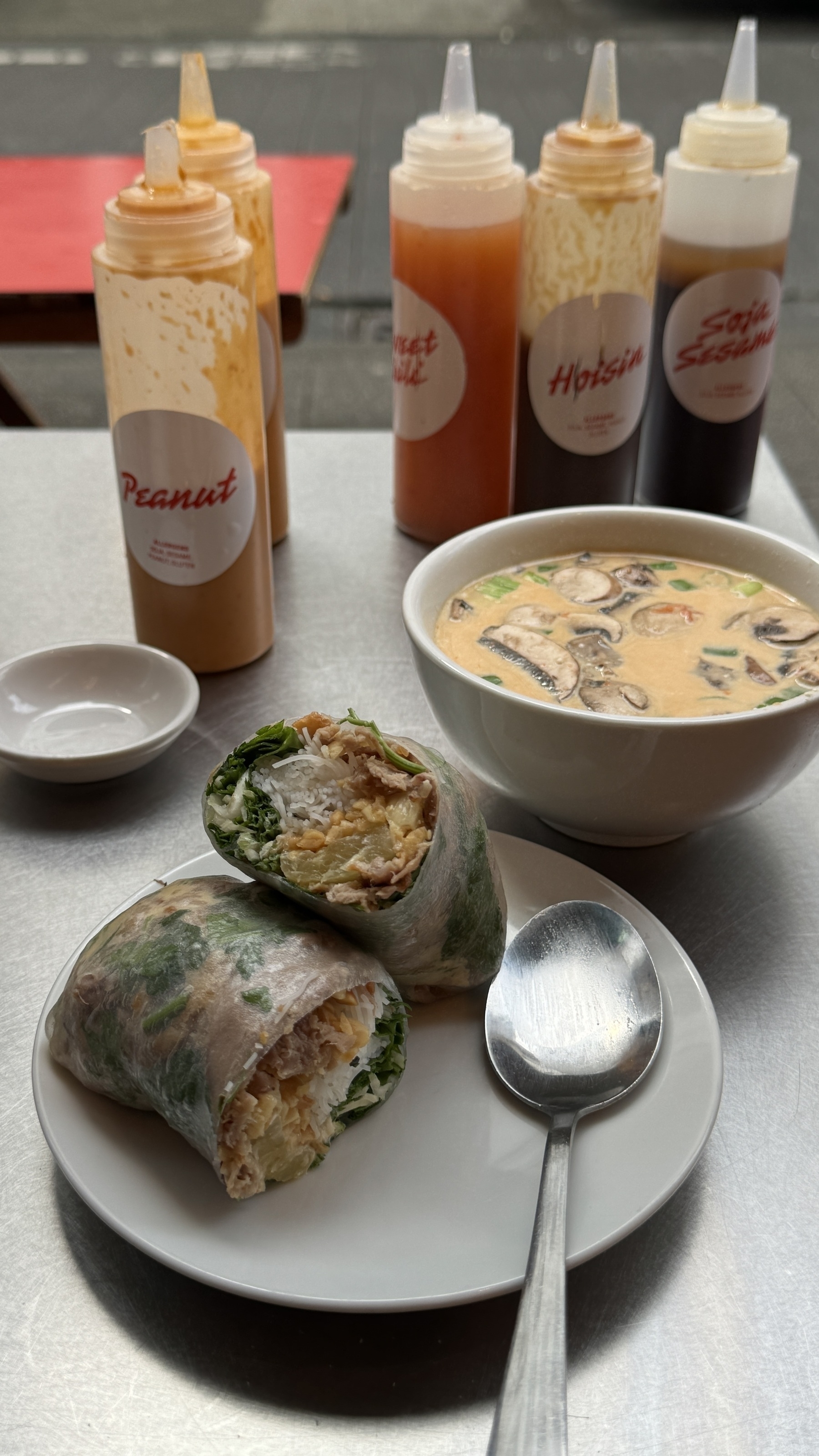 A peanut sauce bottle sits next to a rice-filled spring roll and a bowl of mushroom soup with "Hot Soba Sesame" bottles.
