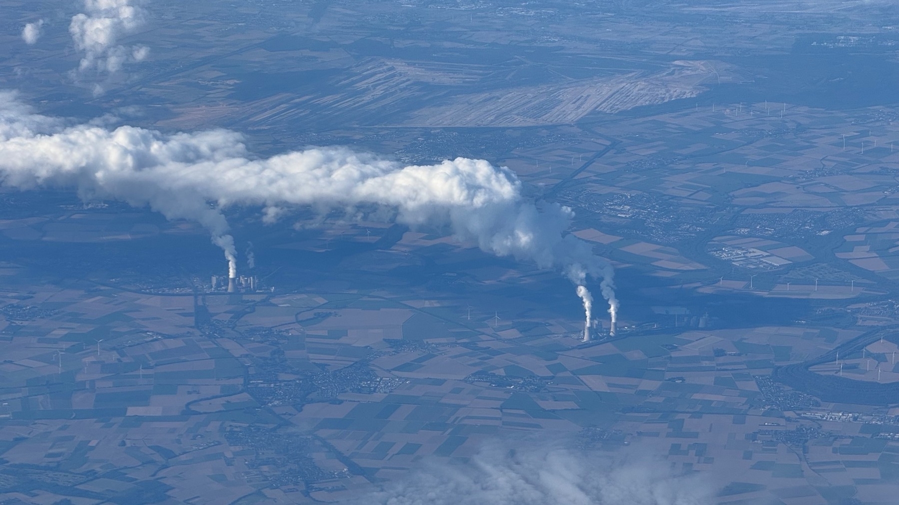 Aerial view shows white clouds rising from industrial smokestacks over a rural landscape with wind turbines.