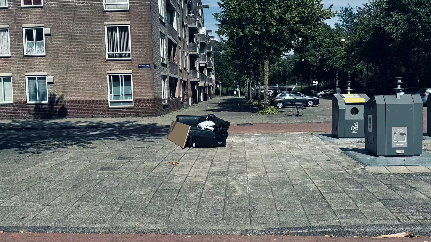 A black sofa sits on a paved street next to two recycling bins with “plastic” labels.