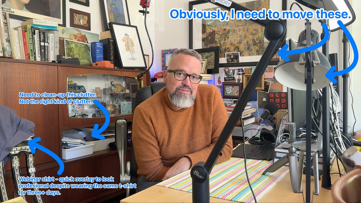 Here's a factual alt-text description for the image: A man wearing a black t-shirt sits at a desk with microphones and books, including "A Walk in the Woods.