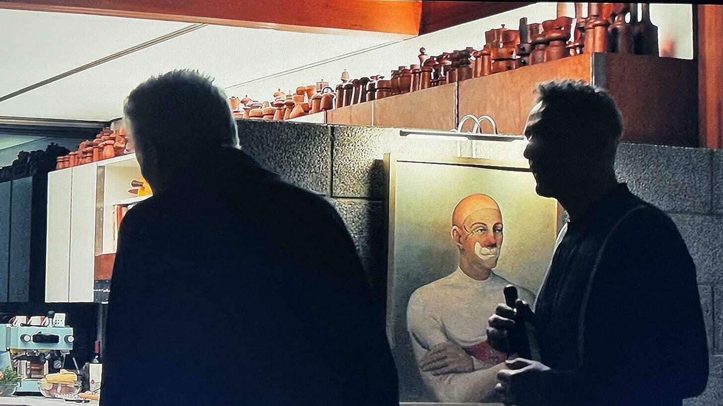 A portrait of Mahatma Gandhi is visible behind two silhouetted figures in a room with a shelf of ceramic objects.