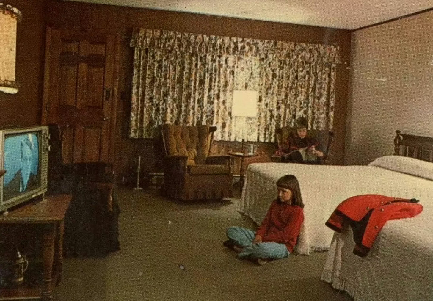 A young girl sits on a floor in a room with a television, armchair, and white bed.