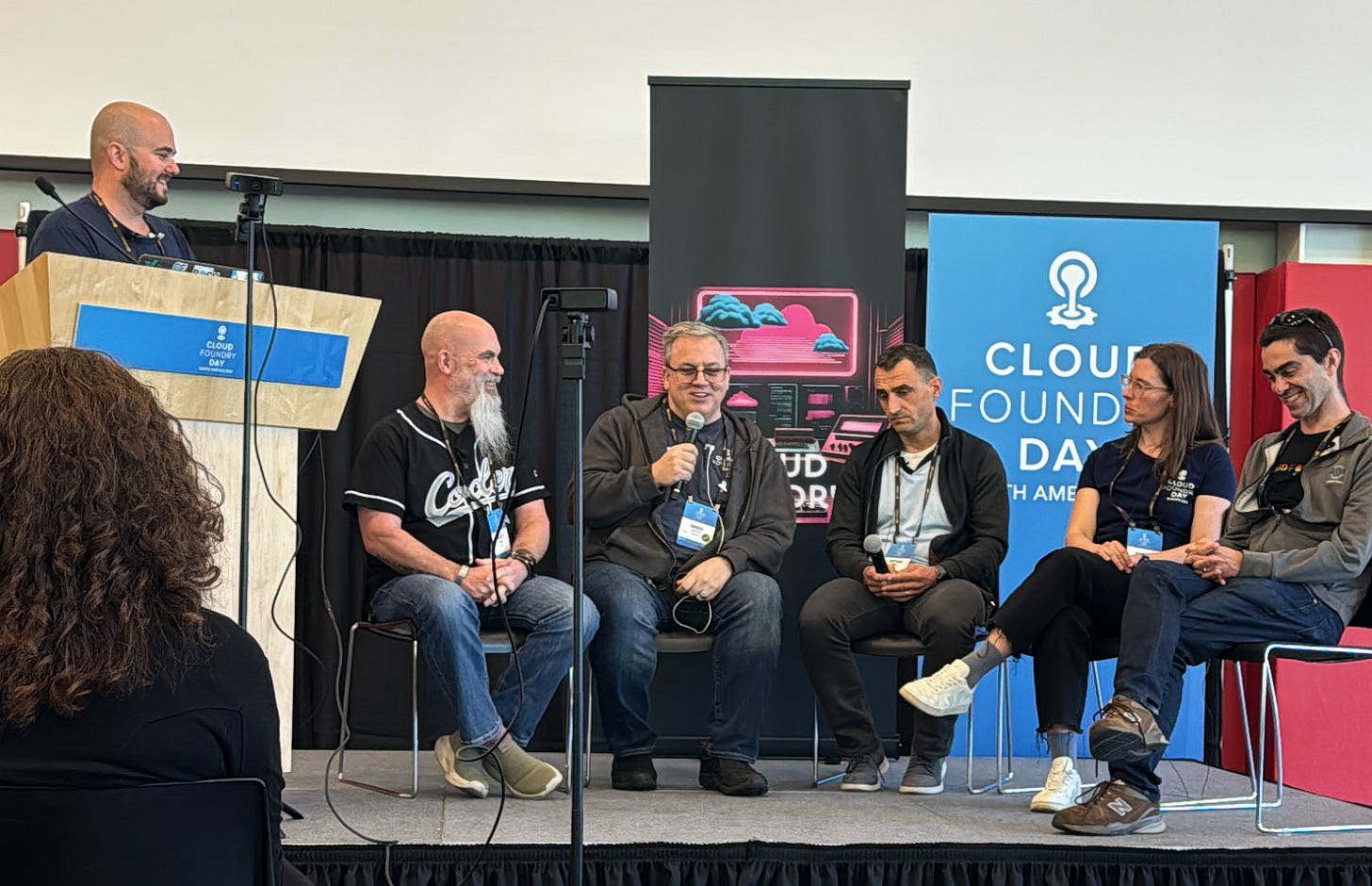 Six people sit on a stage at Cloud Foundry Day, with a blue backdrop displaying "Cloud Foundry Day.