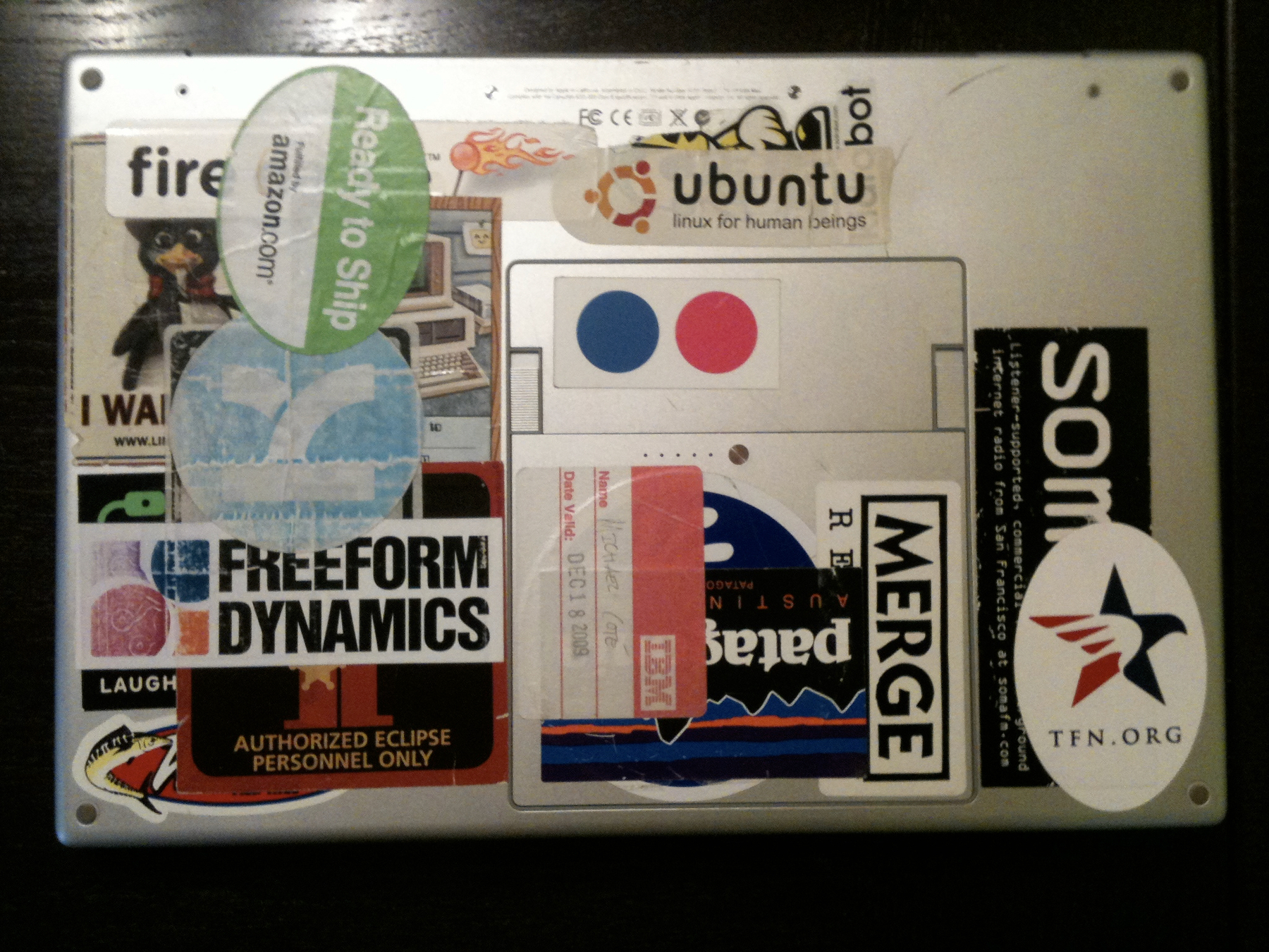 A metal surface is covered with various stickers, including logos for Ready to Ship, Ubuntu, Merge, and others.