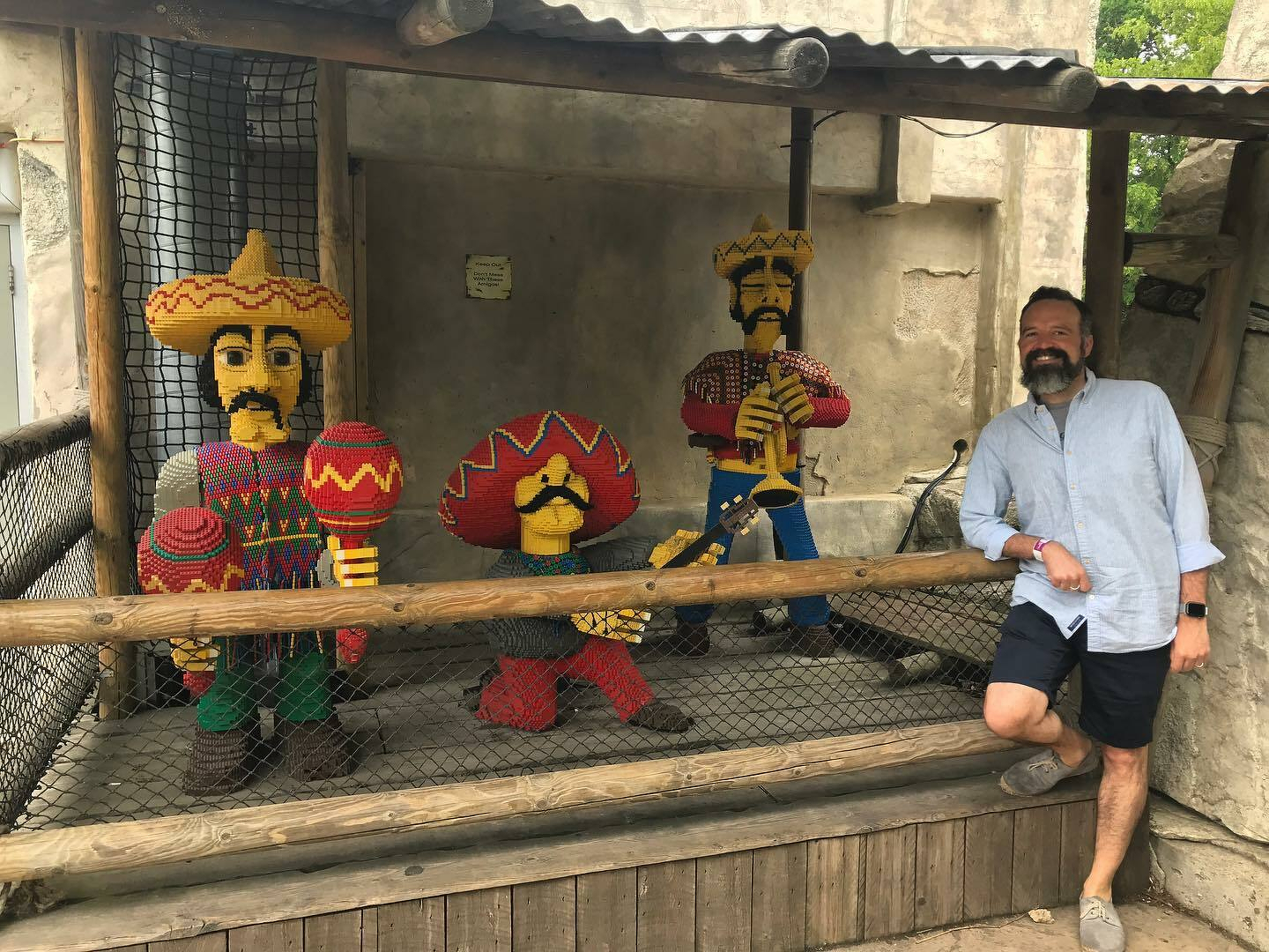 A man stands beside three hand-knitted, miniature Mexican figures behind a wire fence.