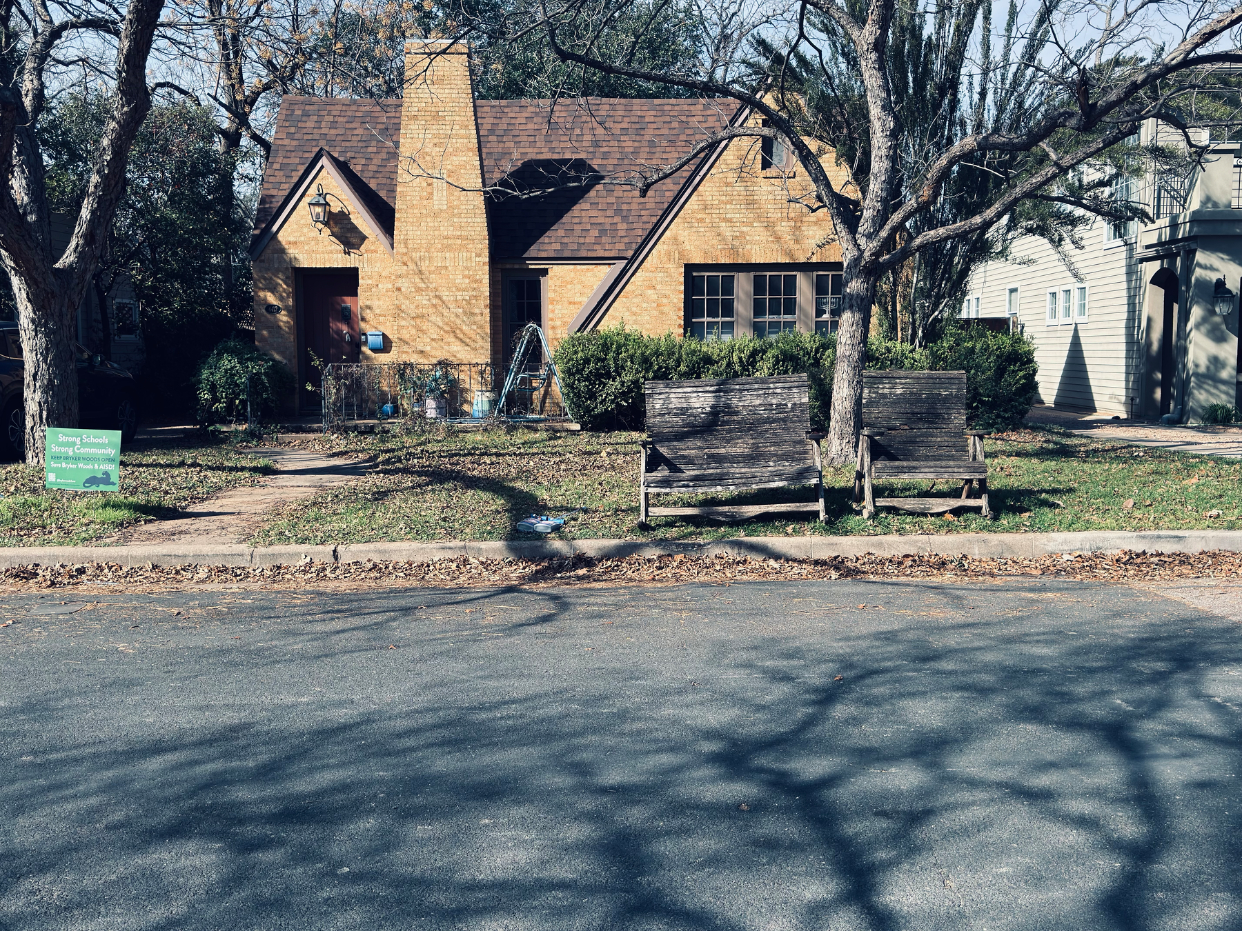 Auto-generated description: A quaint, yellow brick house with a pointed roof and a wooden bench in front is surrounded by bare trees and a lawn with scattered leaves.