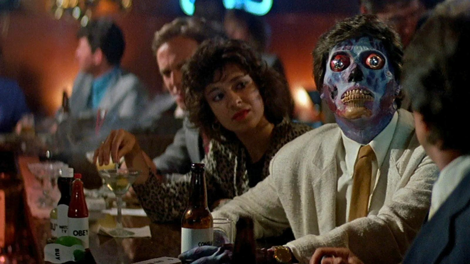 A group of people
  is sitting at a bar, including a person with a half-exposed skeletal face. Still from They Live.