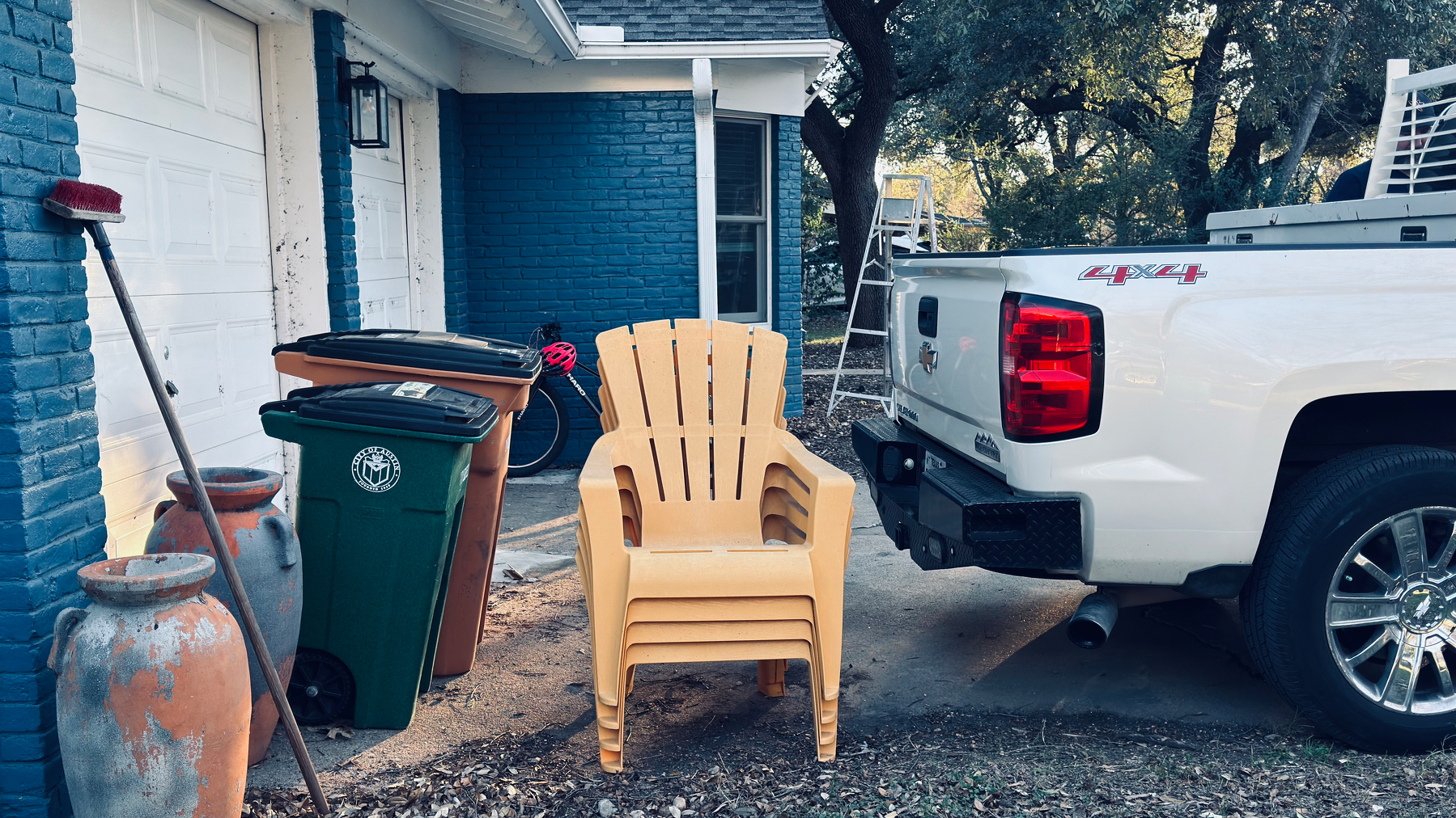 Auto-generated description: A white pickup truck is parked in a driveway next to stacked yellow chairs, trash bins, and large plant pots by a blue house.