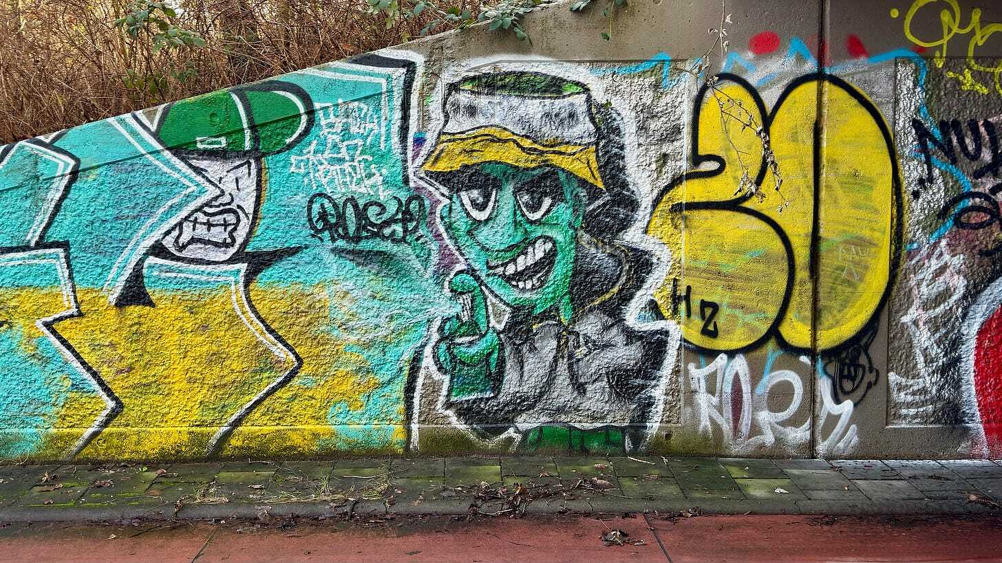 A vibrant graffiti mural features a green-faced character in a bucket hat, alongside various colorful abstract shapes and tags on a concrete wall. Duivendrecht, The Netherlands.