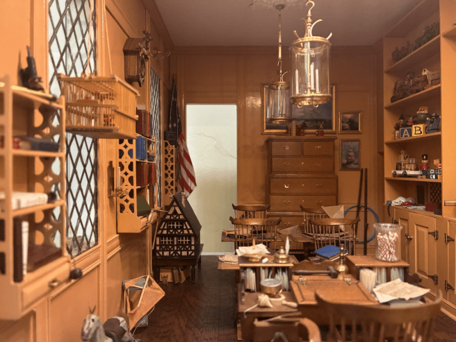 A miniature room features wooden shelves filled with books and toys, with an American flag and the letters "A" and "B" visible.
