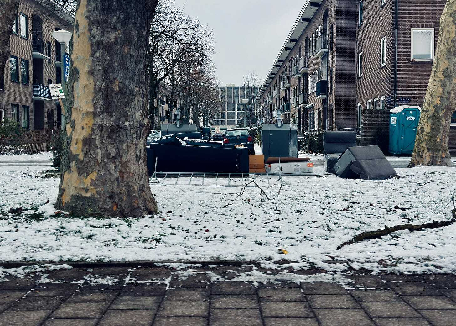 Snow-covered street with a tree, a green portable toilet, and buildings in the background.
