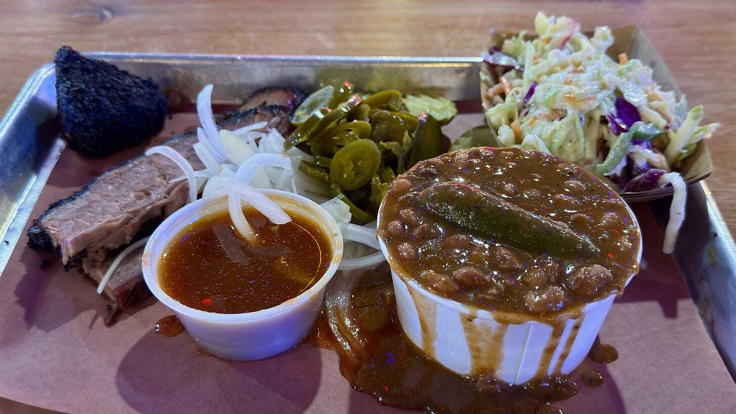 A barbecue tray features smoked brisket, a cup of barbecue sauce, pickled jalapeños, and coleslaw.