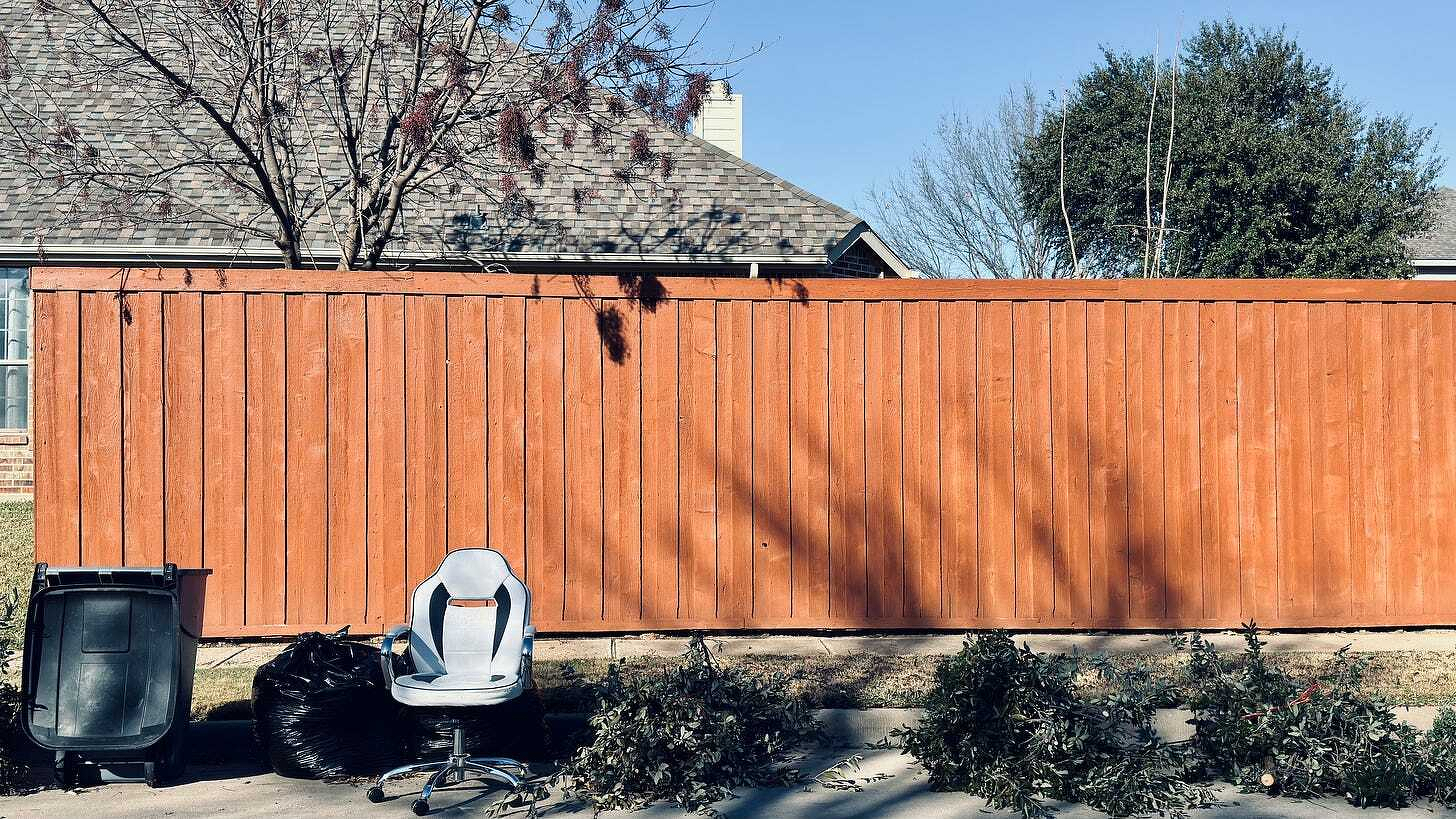 A tall, orange wooden fence runs along a residential street with a dark trash bin and chair nearby.