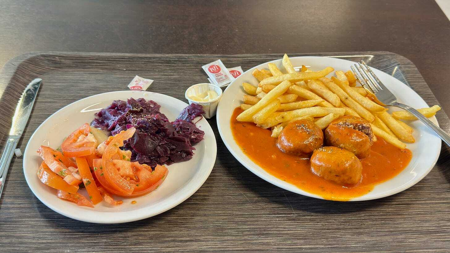 A white tray holds a plate with Swedish meatballs in a red sauce, fries, and a salad with tomatoes.