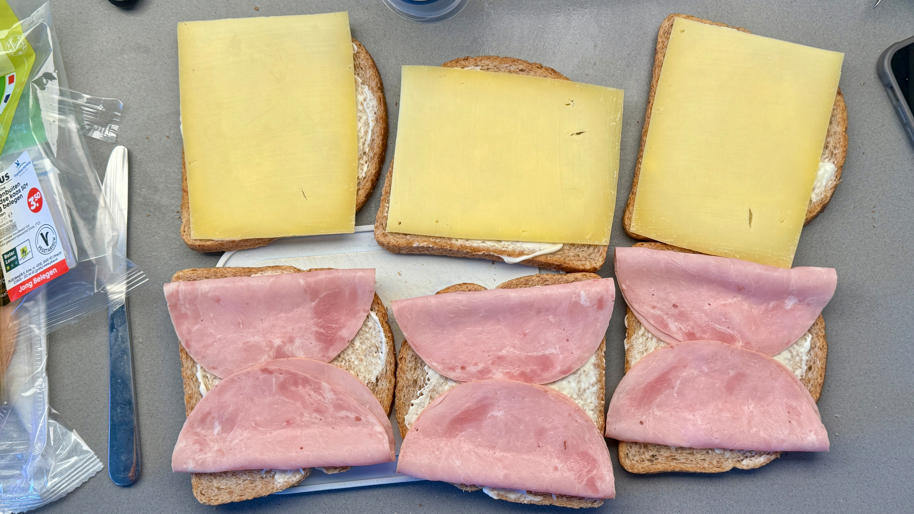 Two sandwiches with ham and cheese are displayed on a gray surface, with packaging visible in the background.