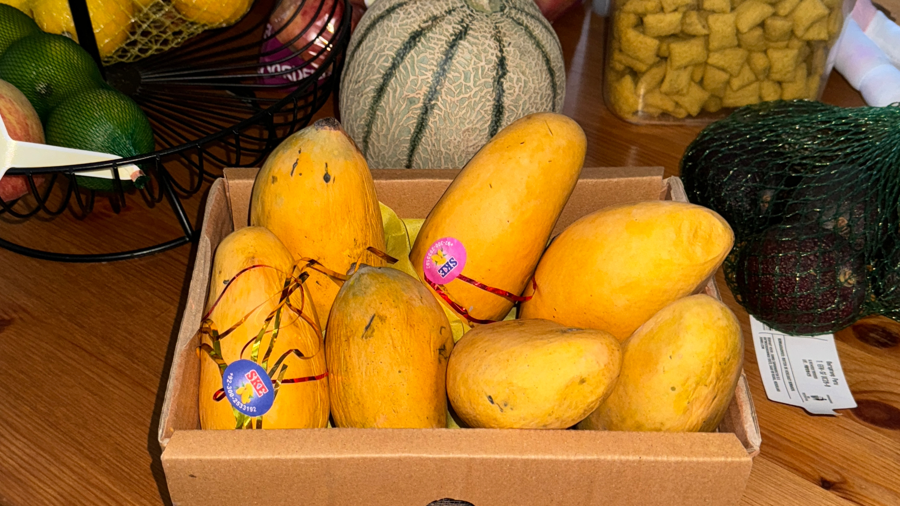 Yellow mangoes in a cardboard box with a SKBS label and a receipt visible.