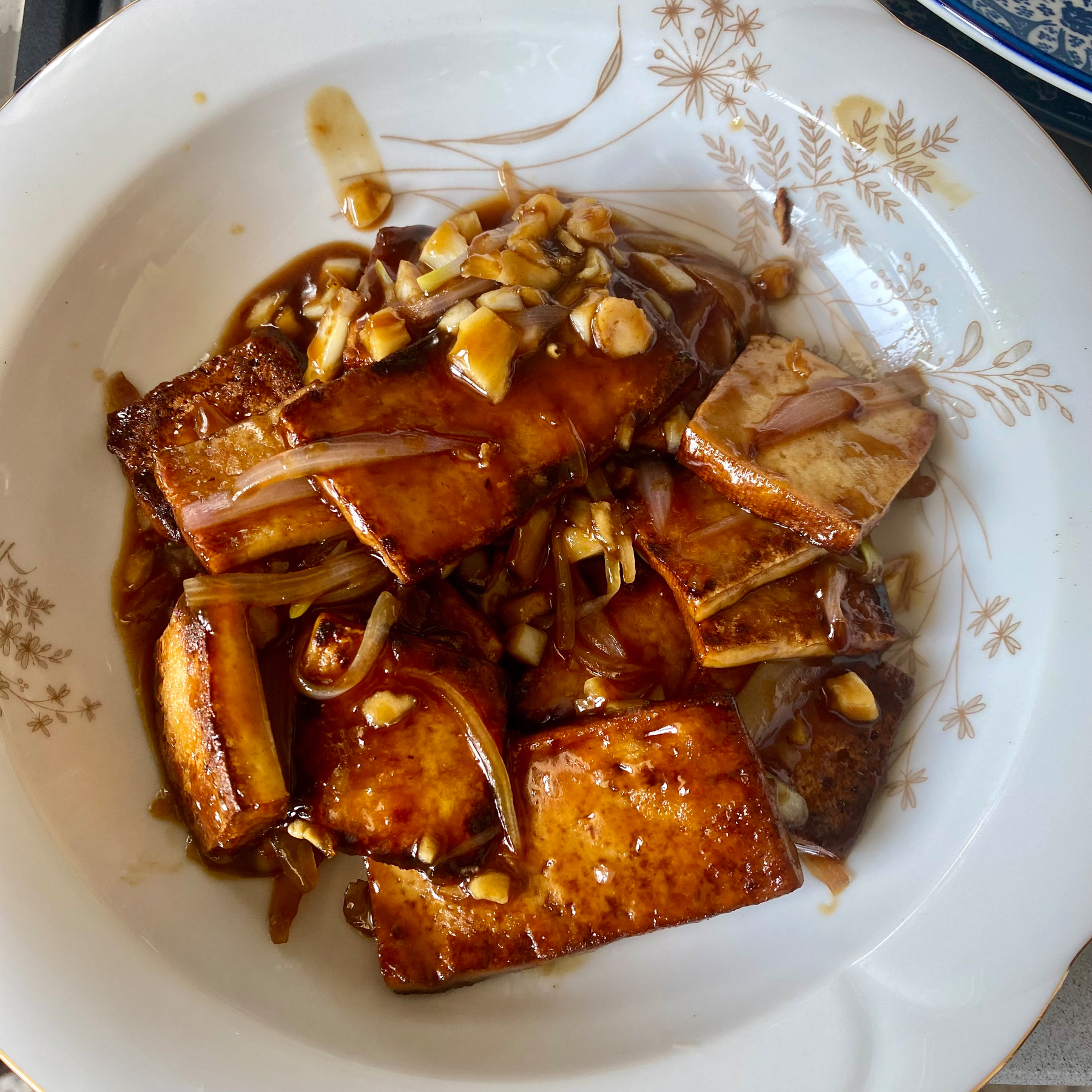 Dark brown tofu cubes with caramelized onions and walnuts are served on a white plate with a floral pattern.