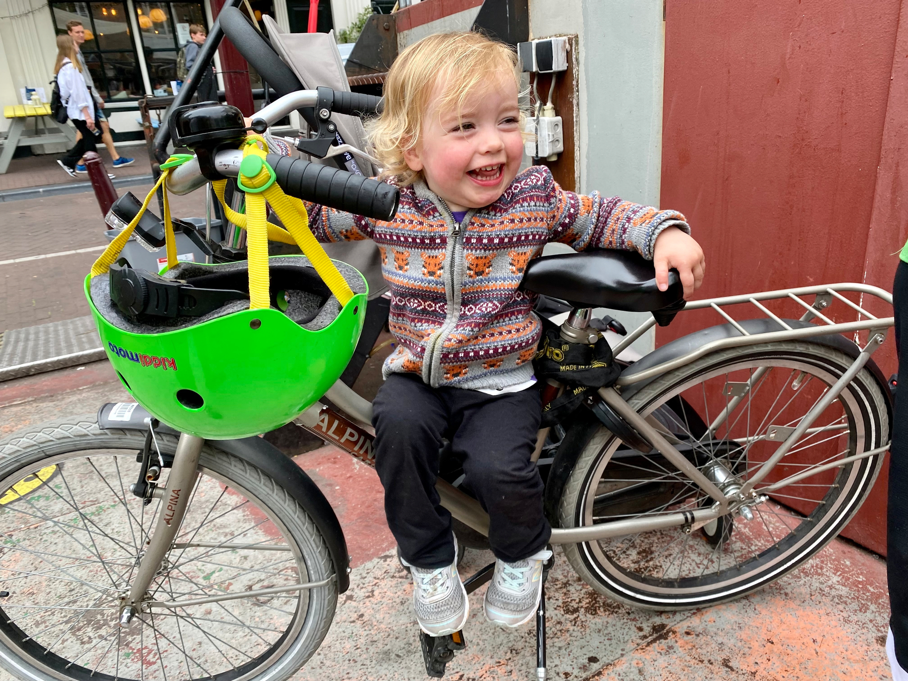 A young child rides a green bicycle with a child seat, wearing a patterned sweater and sneakers.