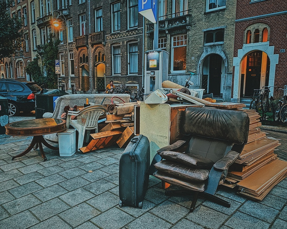 Stacked furniture and a suitcase are arranged on a cobblestone street next to parked bicycles and a building.