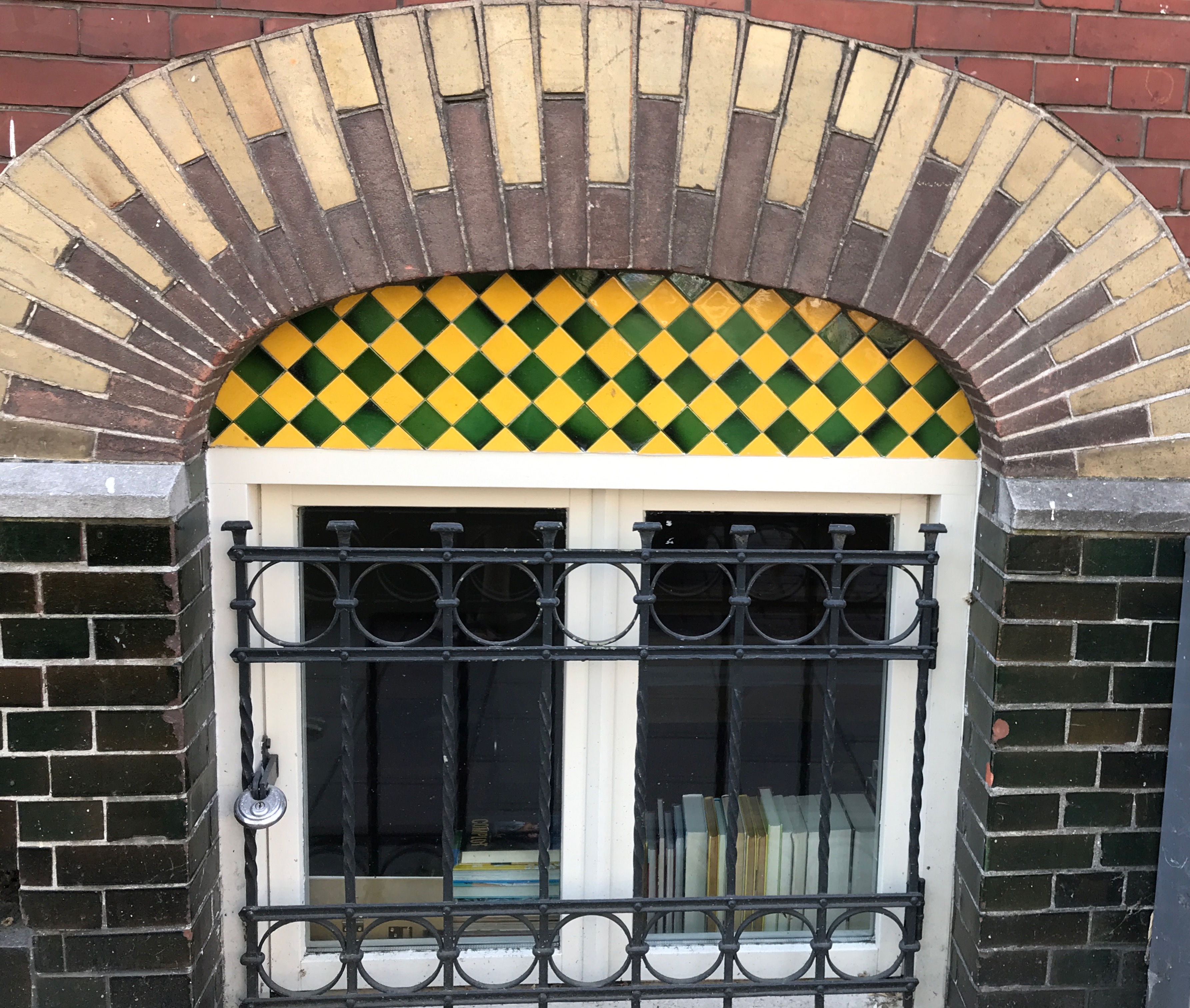 A brick archway with a green and yellow diamond pattern above a window with a black wrought-iron balcony and books inside.