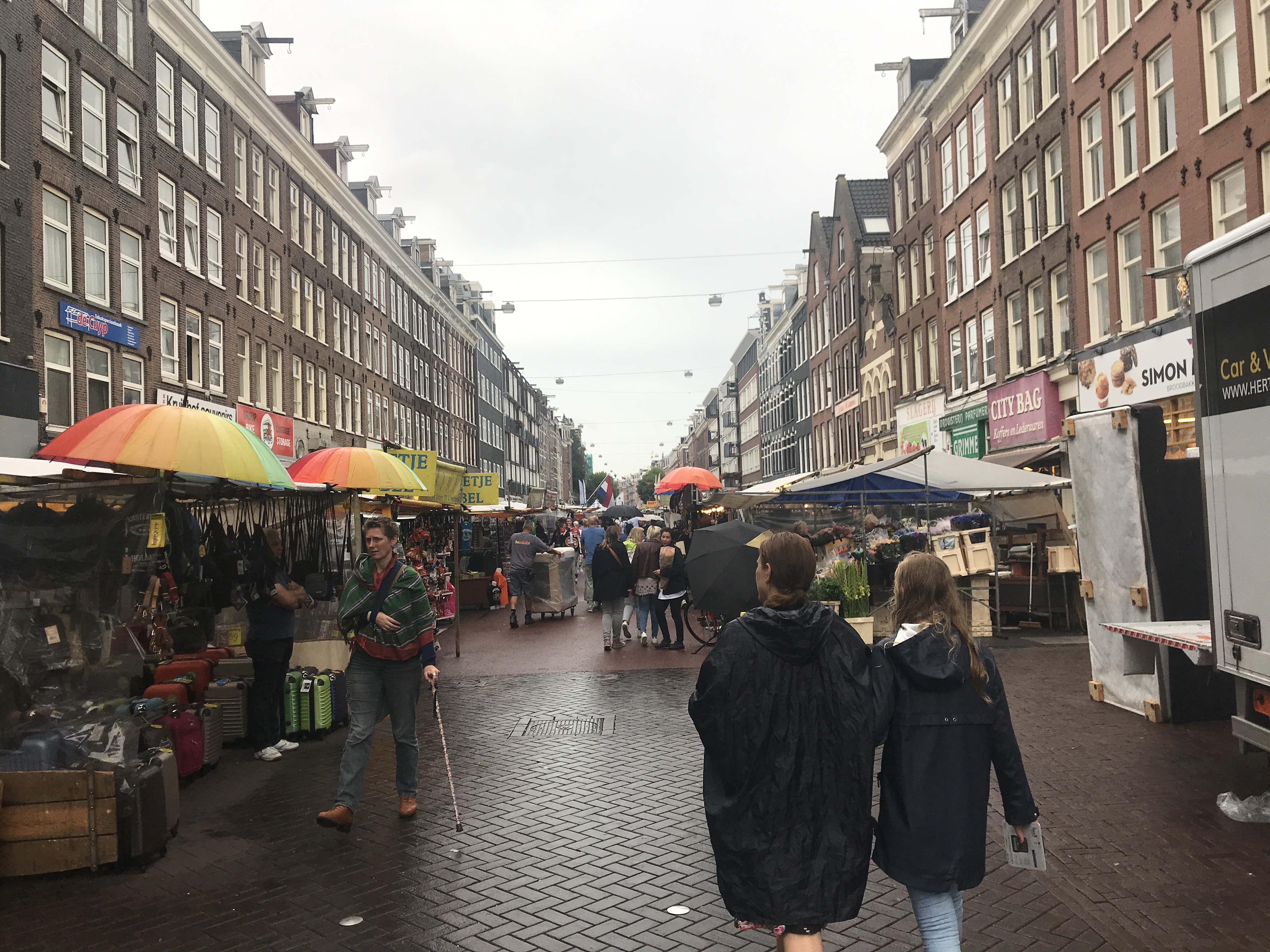 A crowded street in Amsterdam features market stalls under colorful umbrellas and buildings with signage.
