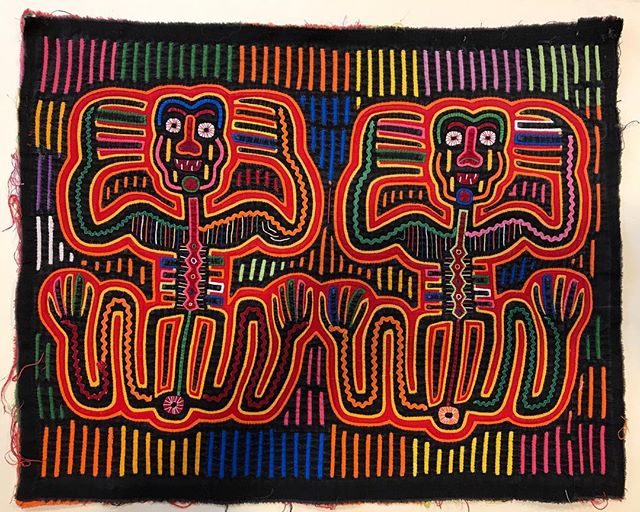 A black textile features two stylized, colorful human figures with intricate embroidery.