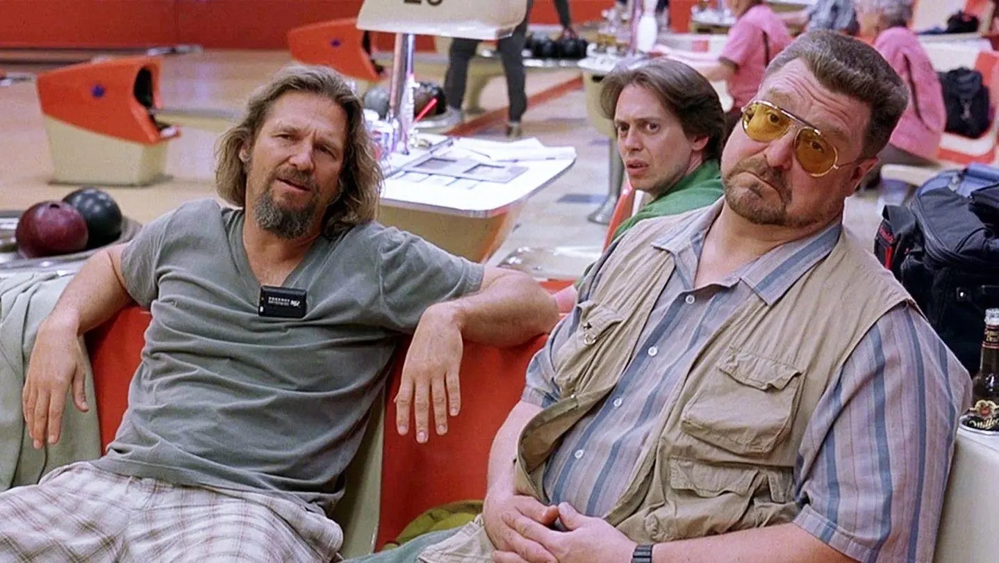 Two men, one with a beard and sunglasses, sit in a bowling alley booth with a beer.