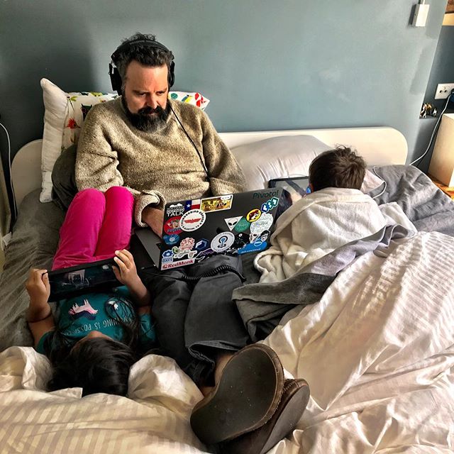 A man with a laptop and headphones is sitting in bed with a child’s feet visible, surrounded by stickers.