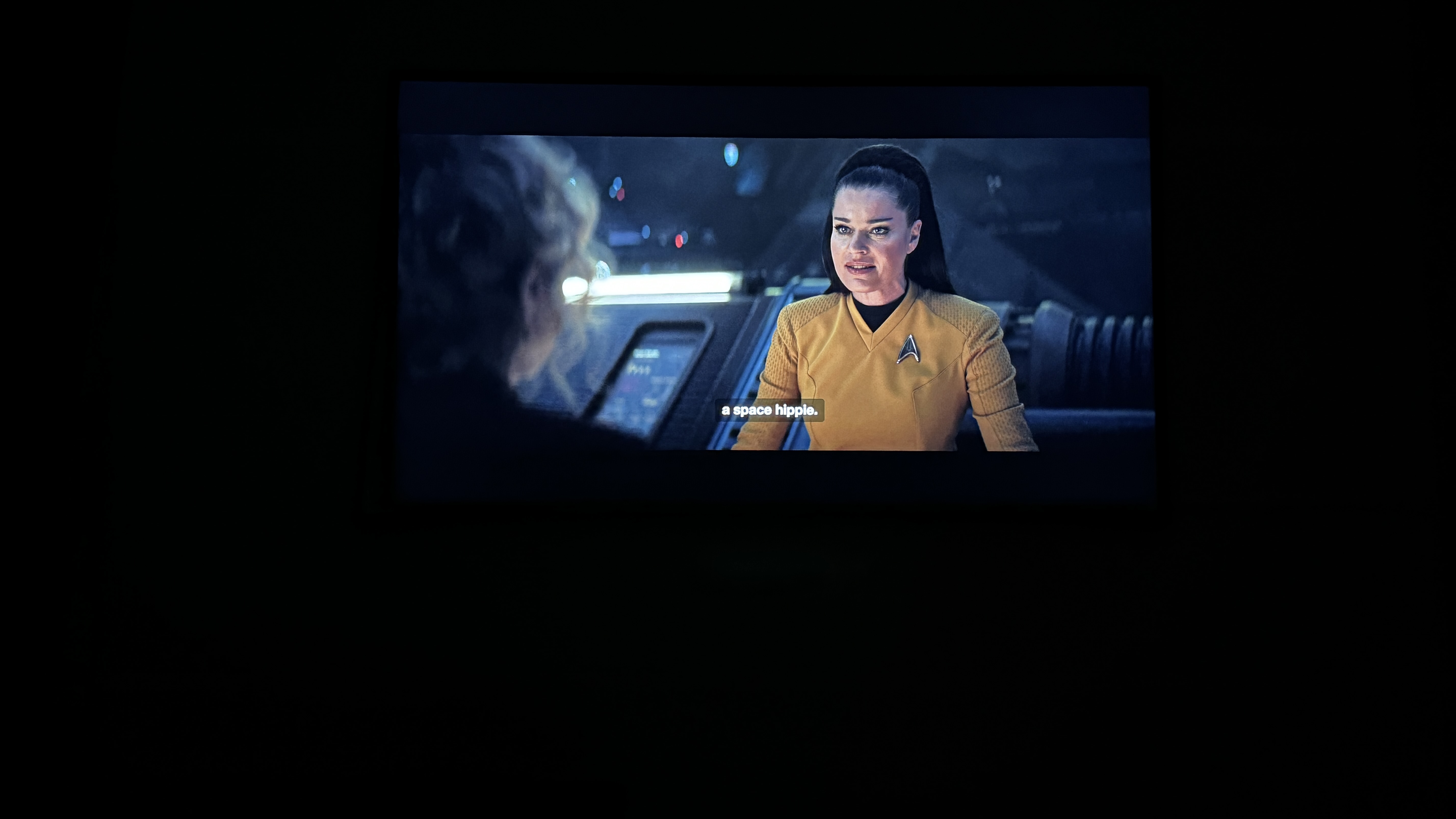 A Starfleet officer sits at a console with a screen displaying data, in a dimly lit space environment.