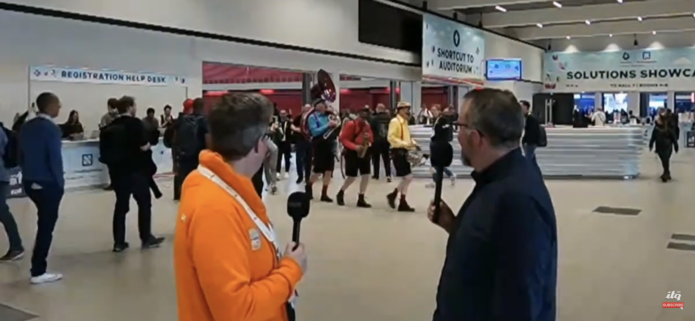 A man in an orange jacket speaks with a man in a black suit at an event registration help desk.