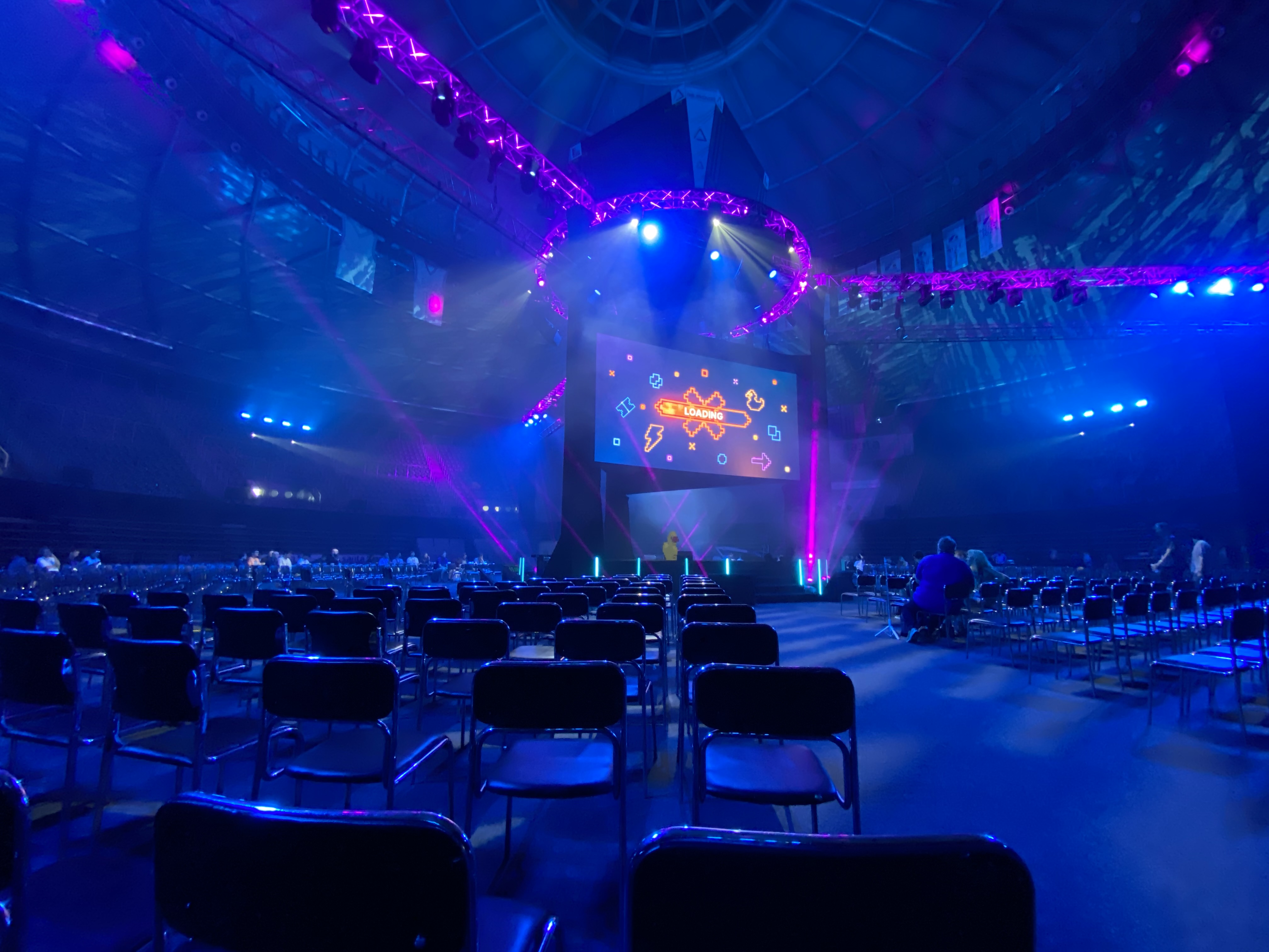 Empty stadium seats are arranged on a stage with a brightly lit screen displaying the word "LOADING".