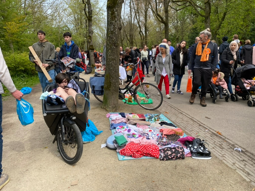 A child relaxes in a bike trailer amid a crowd and a spread of clothing on a path