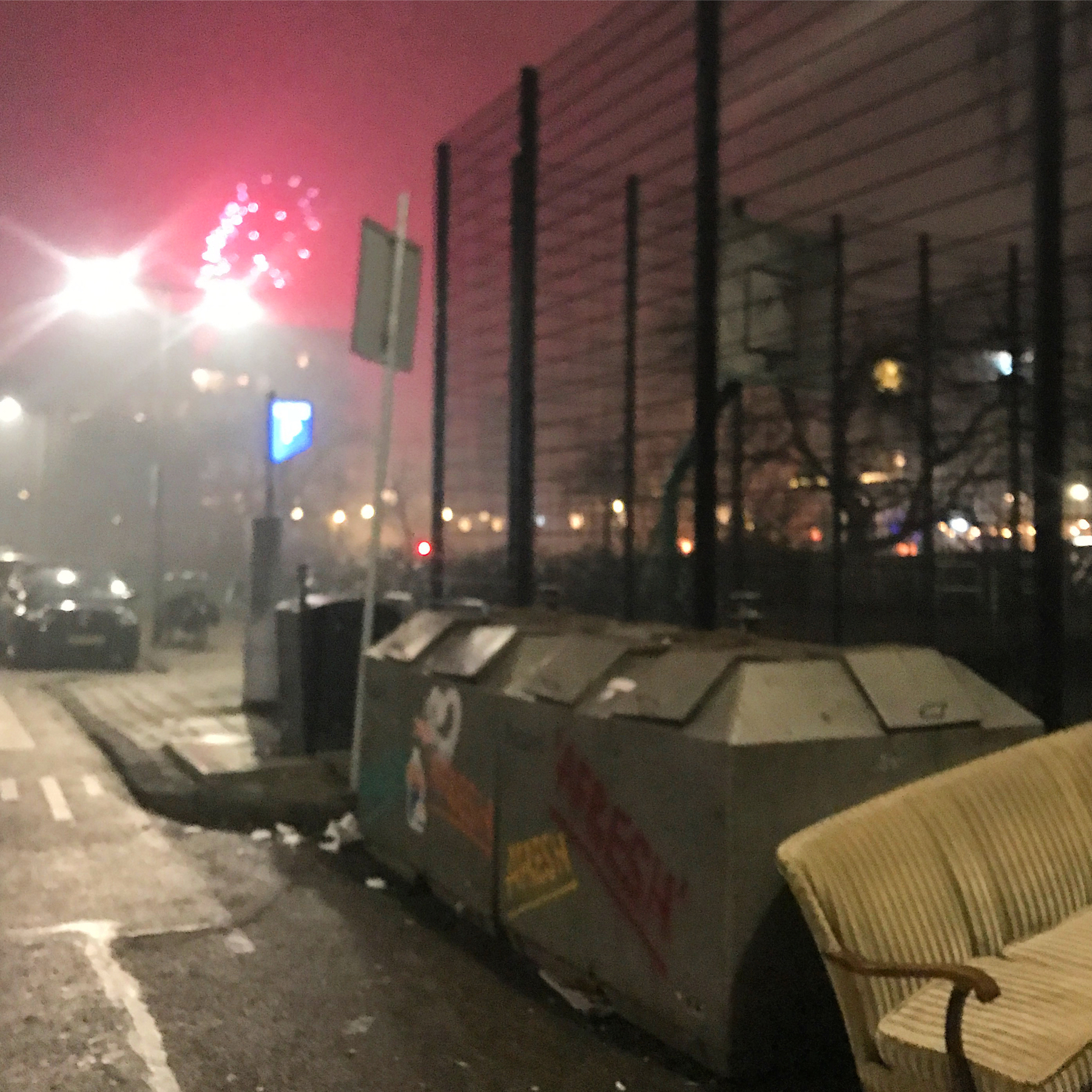 Dumpsters and a bench sit near a road at night under a colorful, overcast sky