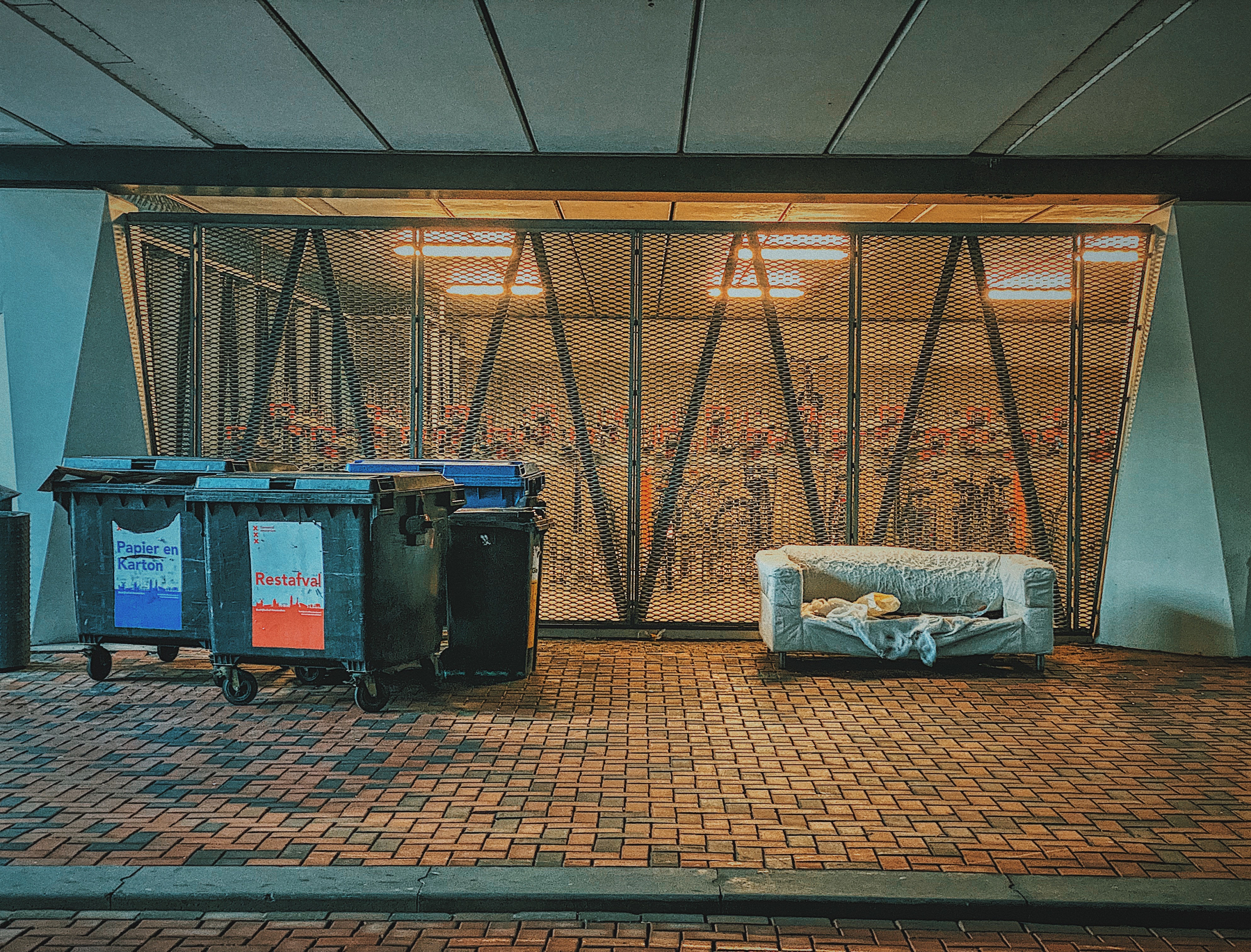 Dumpsters and a discarded sofa sit before a brick wall with a metal grate