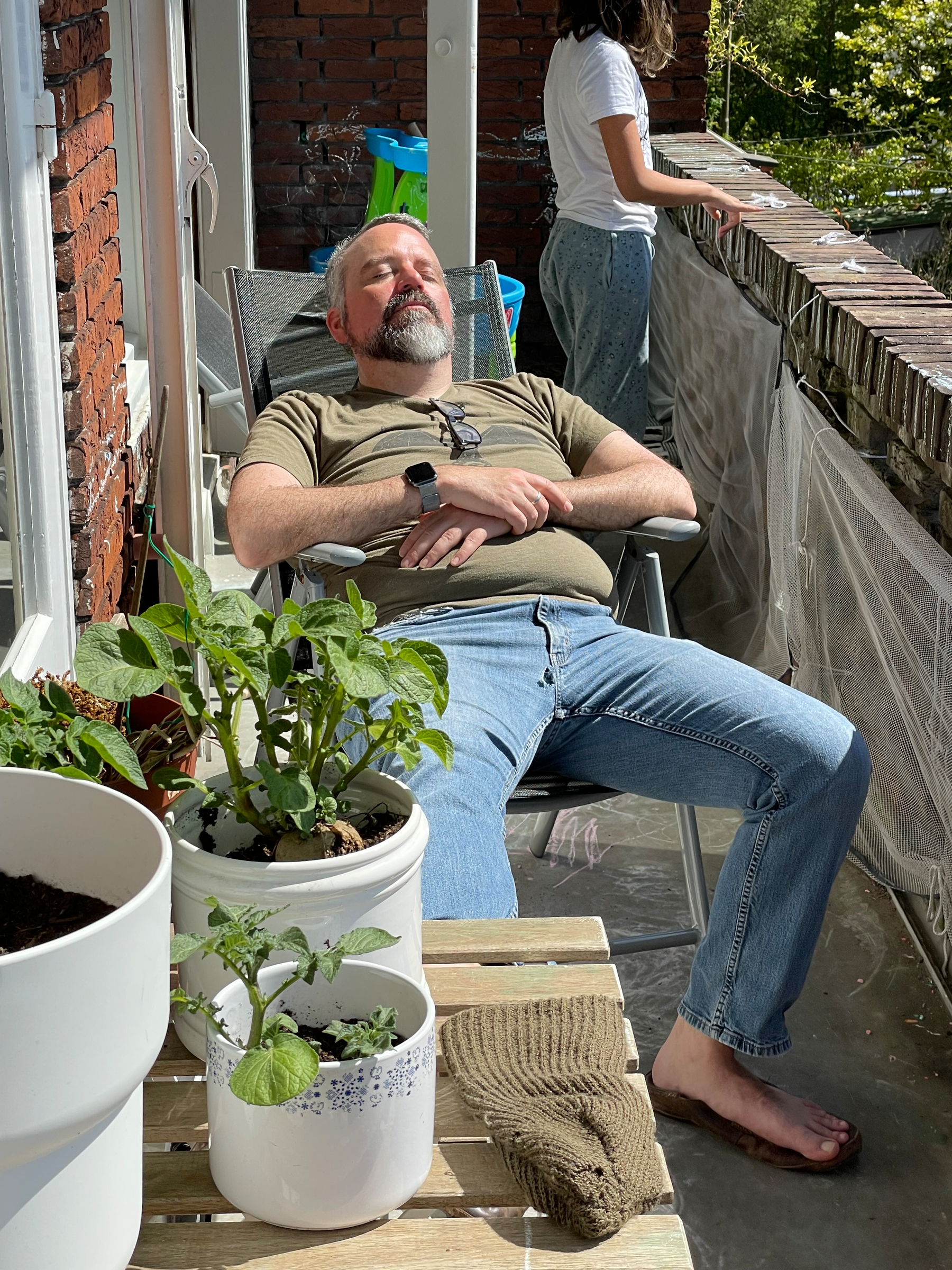 Man relaxes on a balcony, barefoot, with potted plants and a view