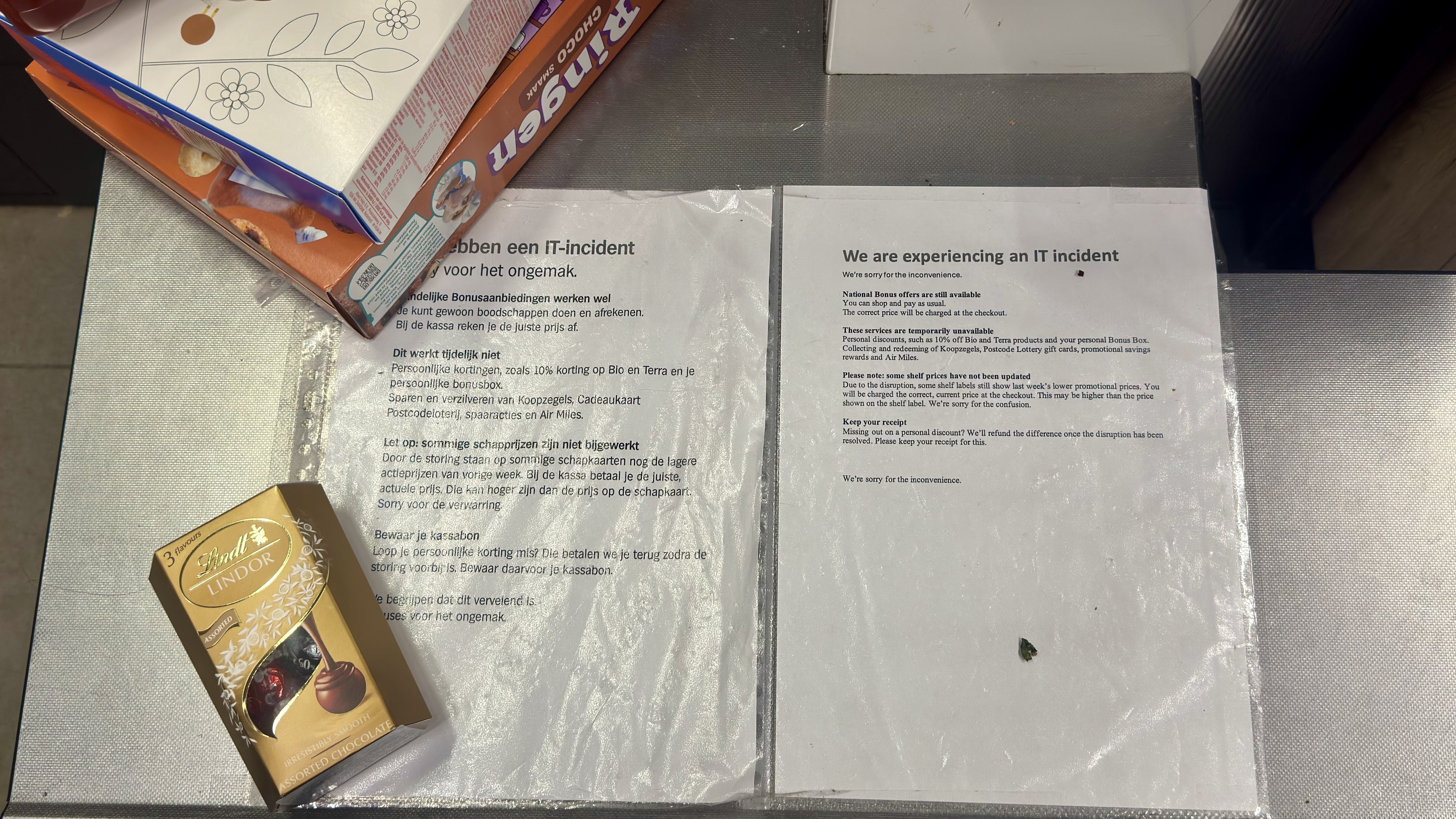 Printed notices in Dutch and English taped to a supermarket checkout counter, announcing an IT incident, next to a box of Lindt chocolates and a stack of snack boxes.
