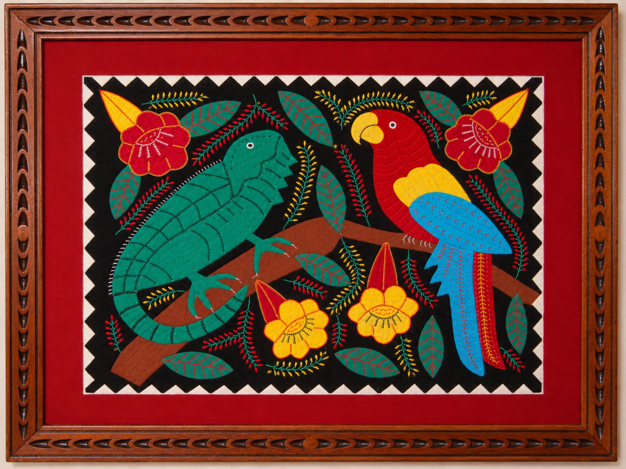 Framed textile artwork depicting a green iguana and a red macaw perched together on a branch among flowers and leaves.