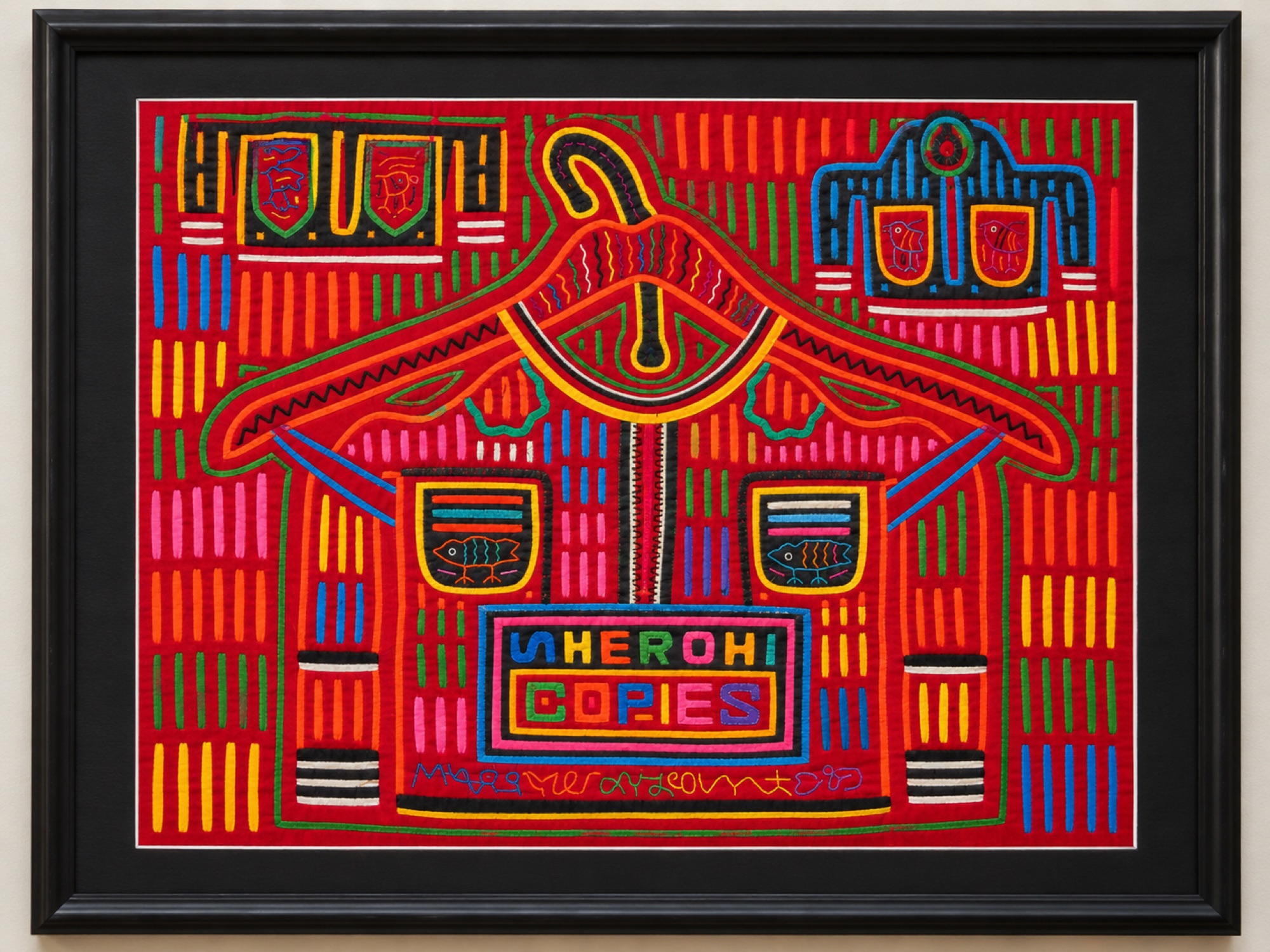 Framed red textile panel displaying a jacket on a clothes hanger with fish in the pockets and decorative lettering.