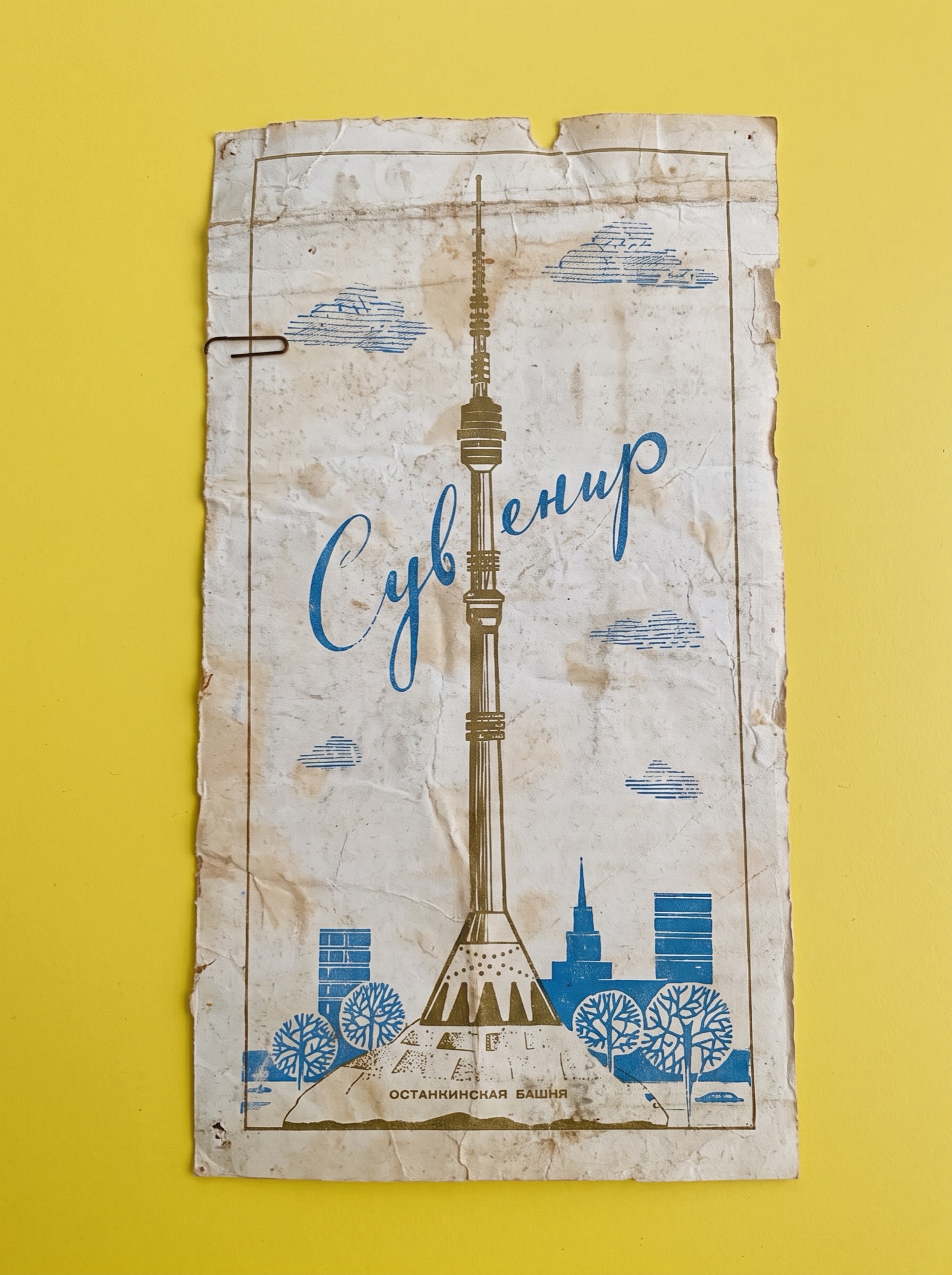 A worn Soviet-era paper souvenir bag on a yellow background, printed in gold with Moscow's Ostankino TV tower and the blue cursive word Сувенир.
