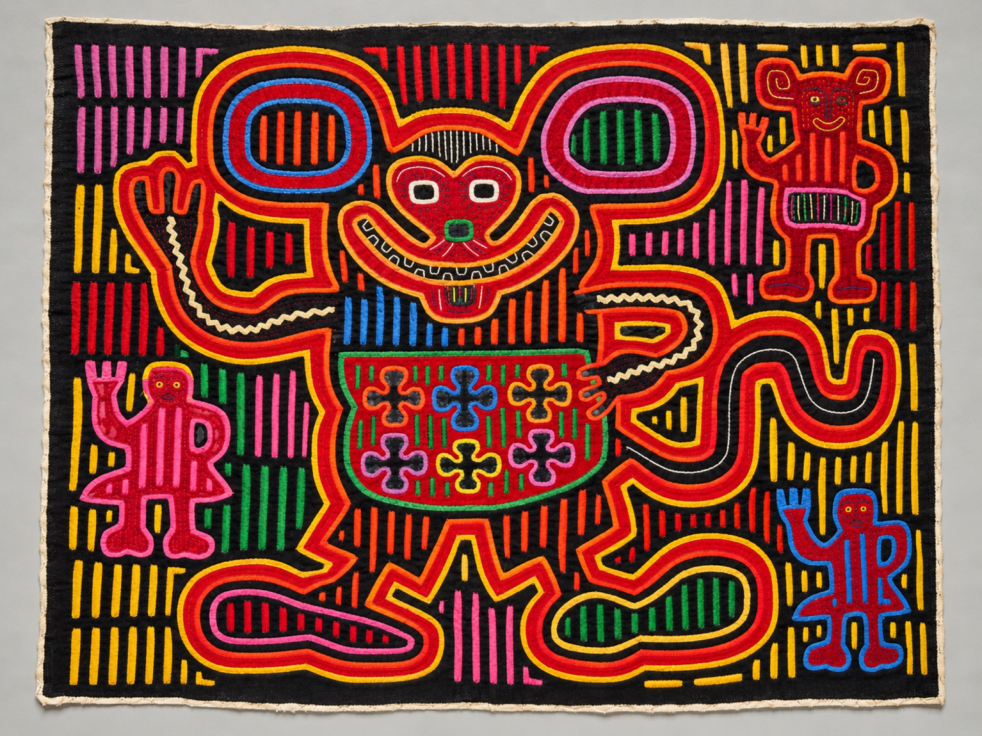 Textile panel showing a large smiling creature with round ears surrounded by smaller waving figures on a striped background.